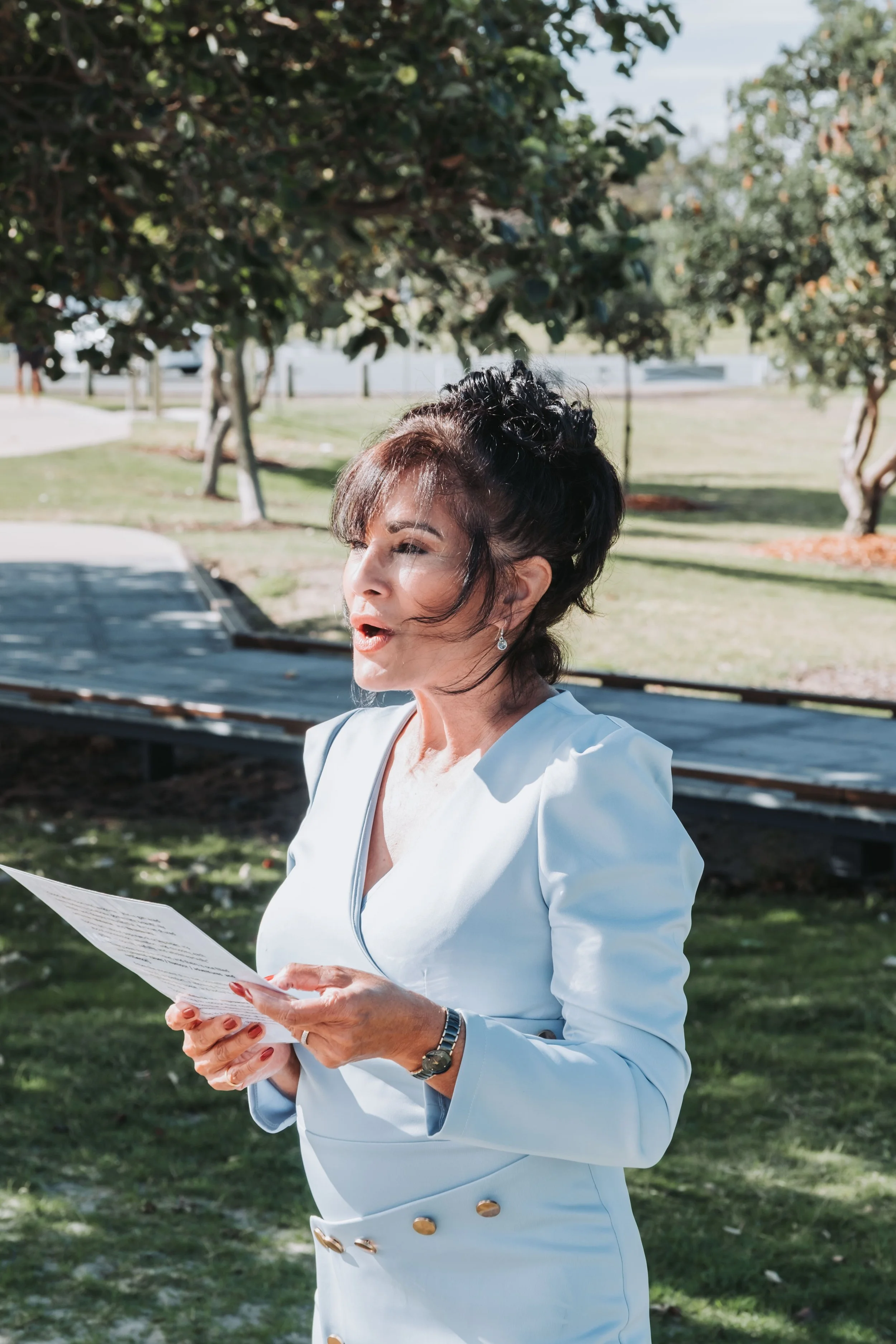 A woman wearing a white blazer is outdoors, reading from a piece of paper, with trees and a park in the background.