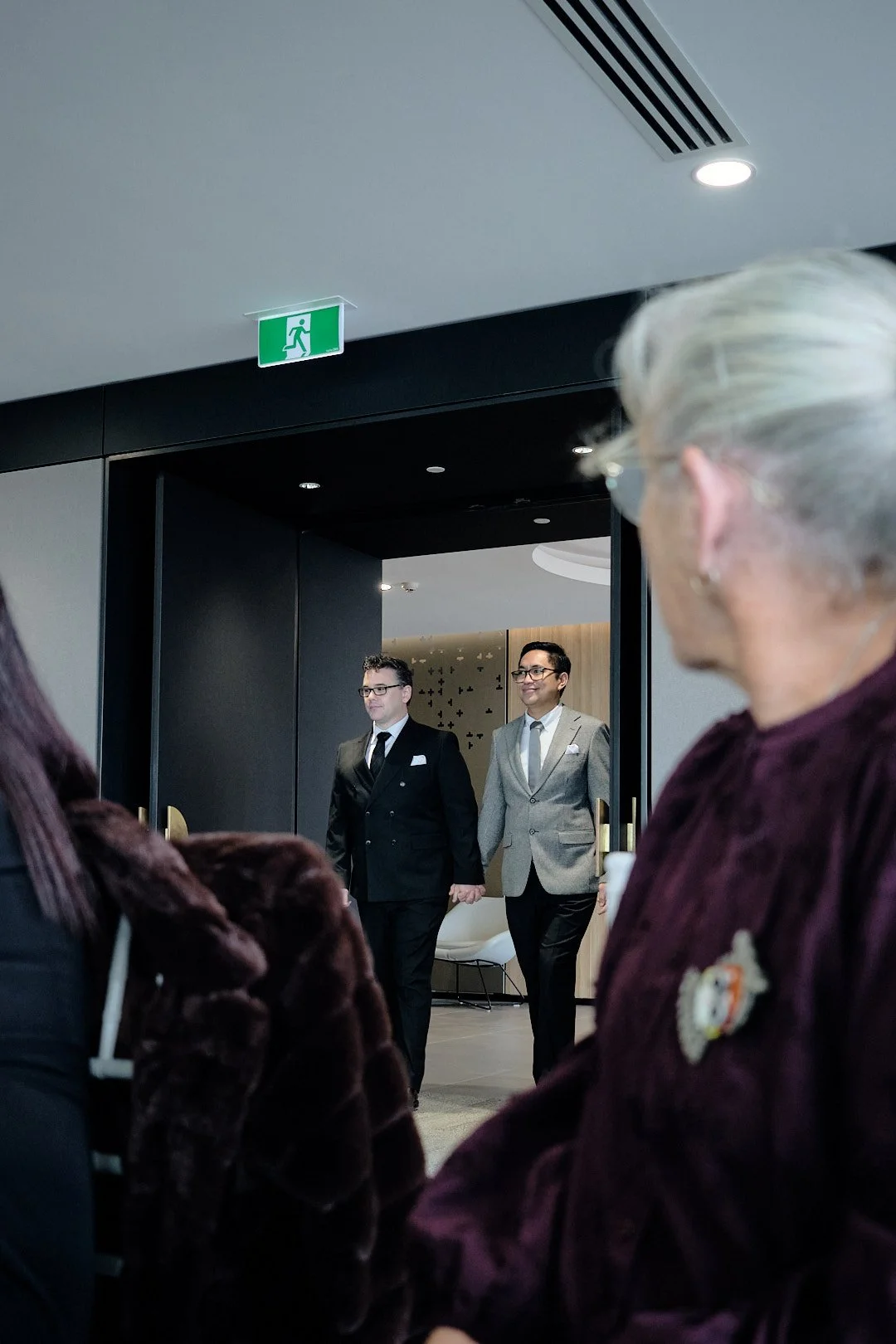 Two men in suits walking through a doorway in a modern indoor space, with a green emergency exit sign above the door and several seated people observing.