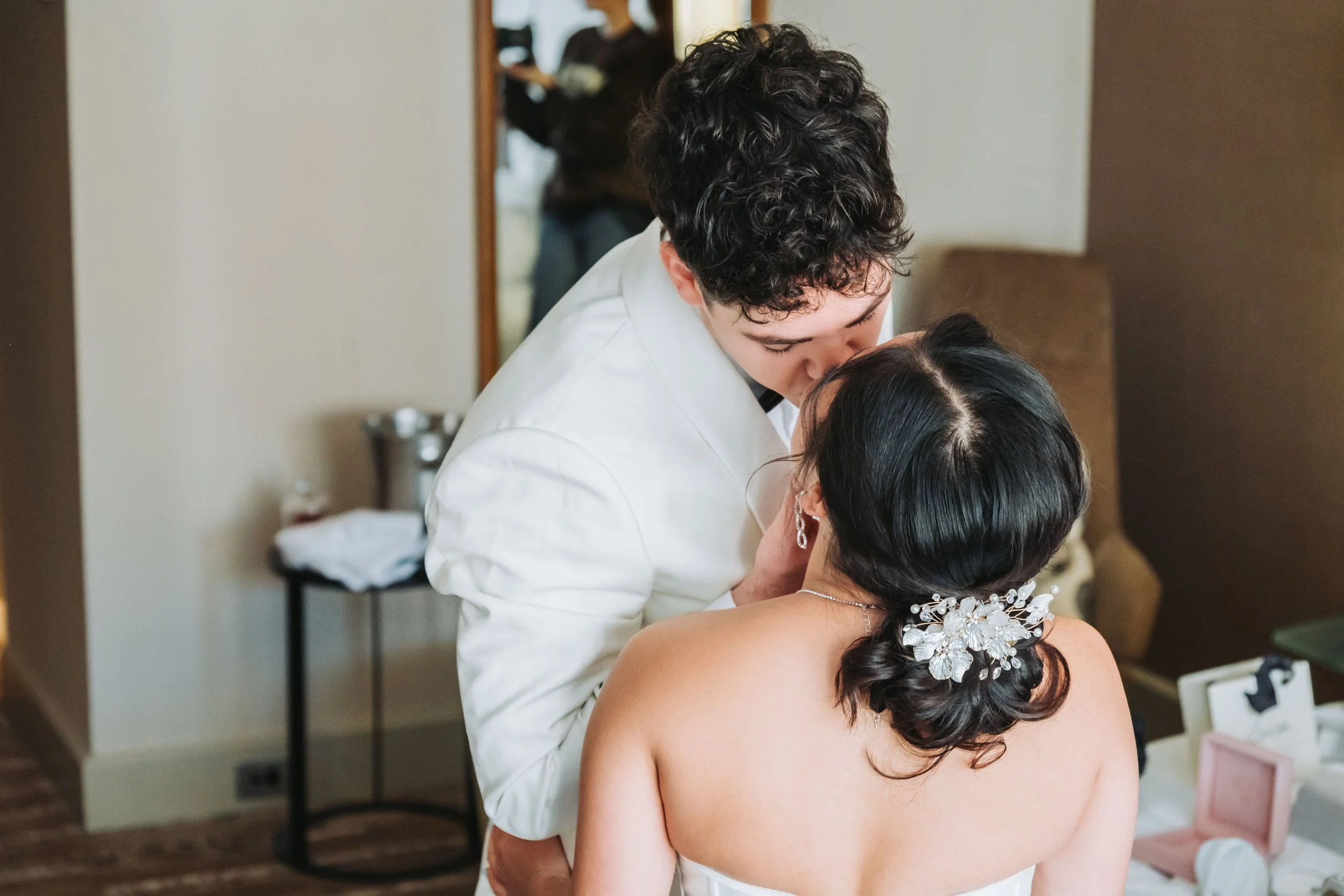 A man in a white suit leans over a woman with long dark hair, wearing a white dress and floral hair accessory, as they share a kiss in a room with a mirror and makeup items in the background.