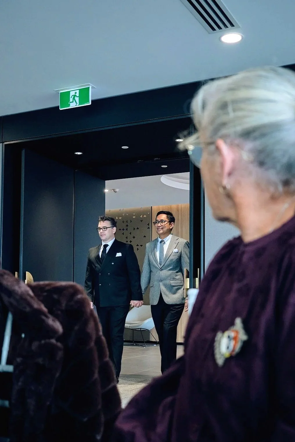 Two grooms in suits hold hands entering a room as guests in burgundy fur coats look on at a wedding.