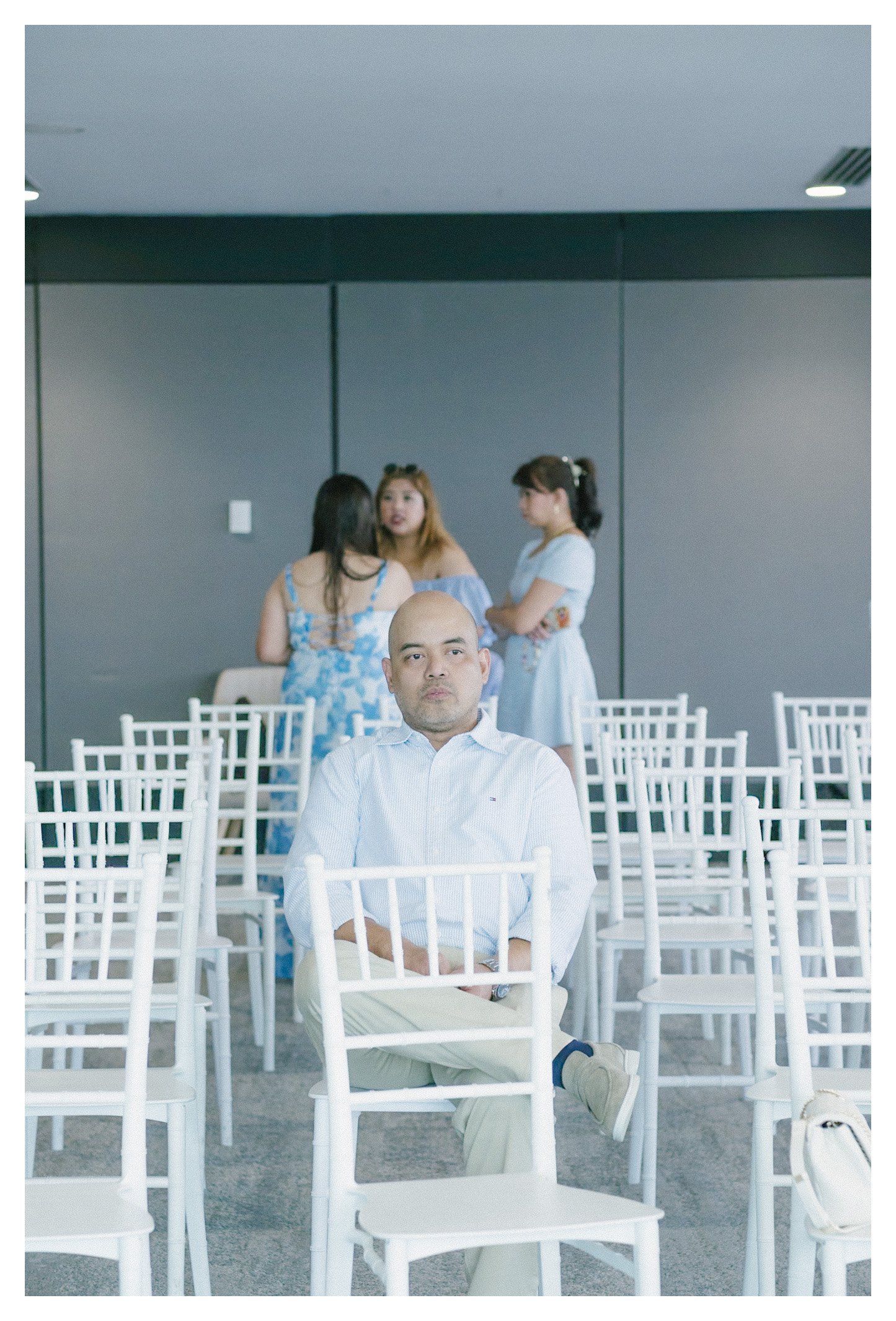 A man wearing a white shirt and beige pants is sitting on a white chair in an empty, modern event space with white chairs. Four women are standing and talking in the background near a gray wall, dressed in casual and semi-formal attire.