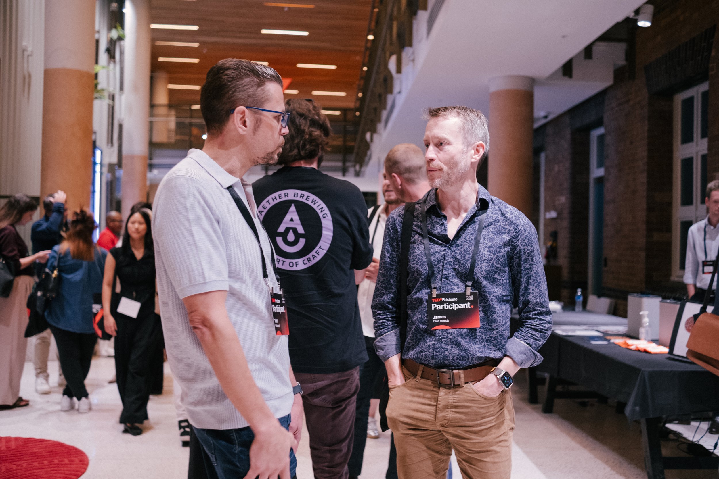 Two men engaged in conversation at a conference or event, wearing name tags that identify one as James Chin Moody, with others visible in the background.