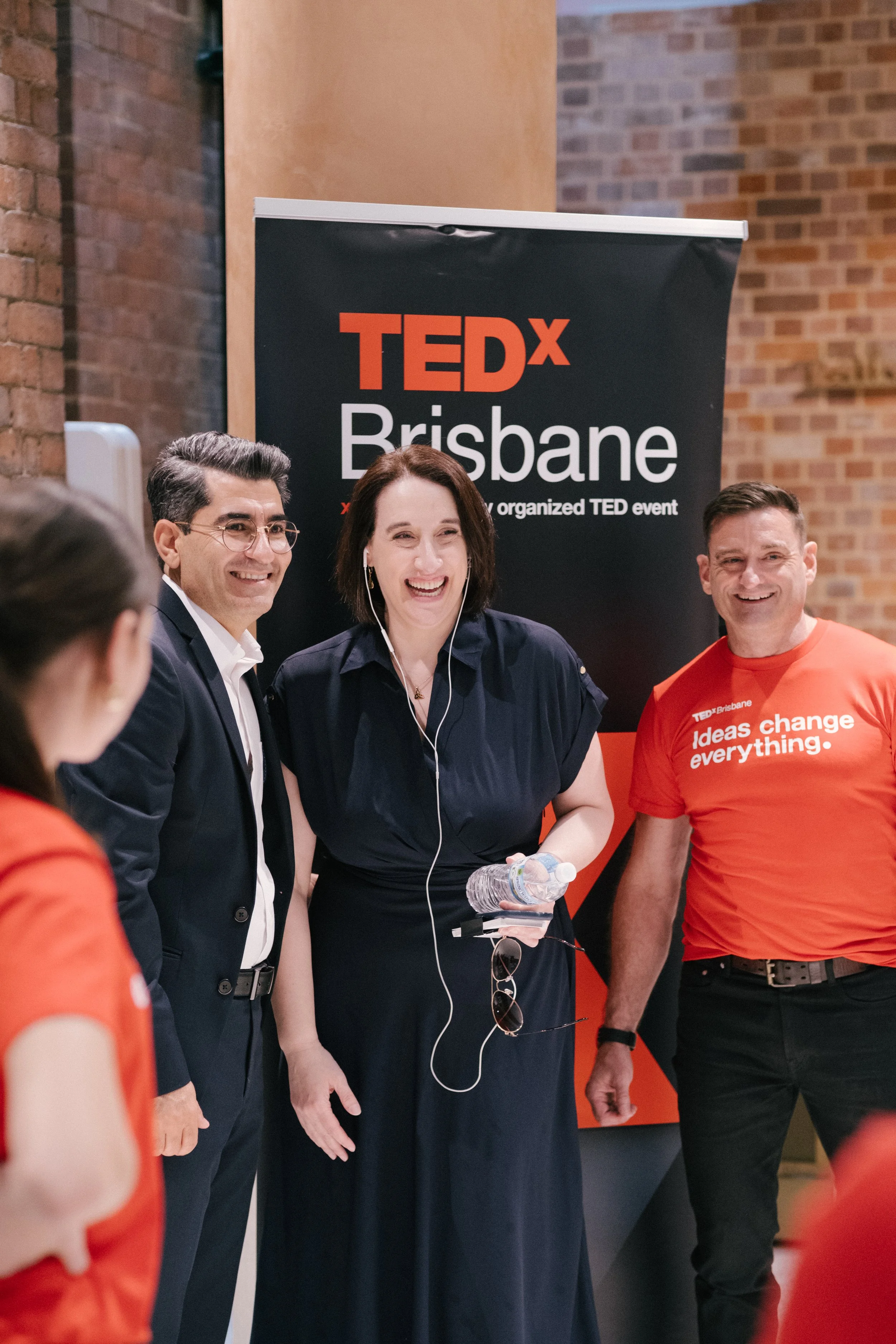 A group of people at a TEDx event in Brisbane, smiling and standing near a black and red TEDx Brisbane banner. One woman is holding a water bottle and wearing earphones.
