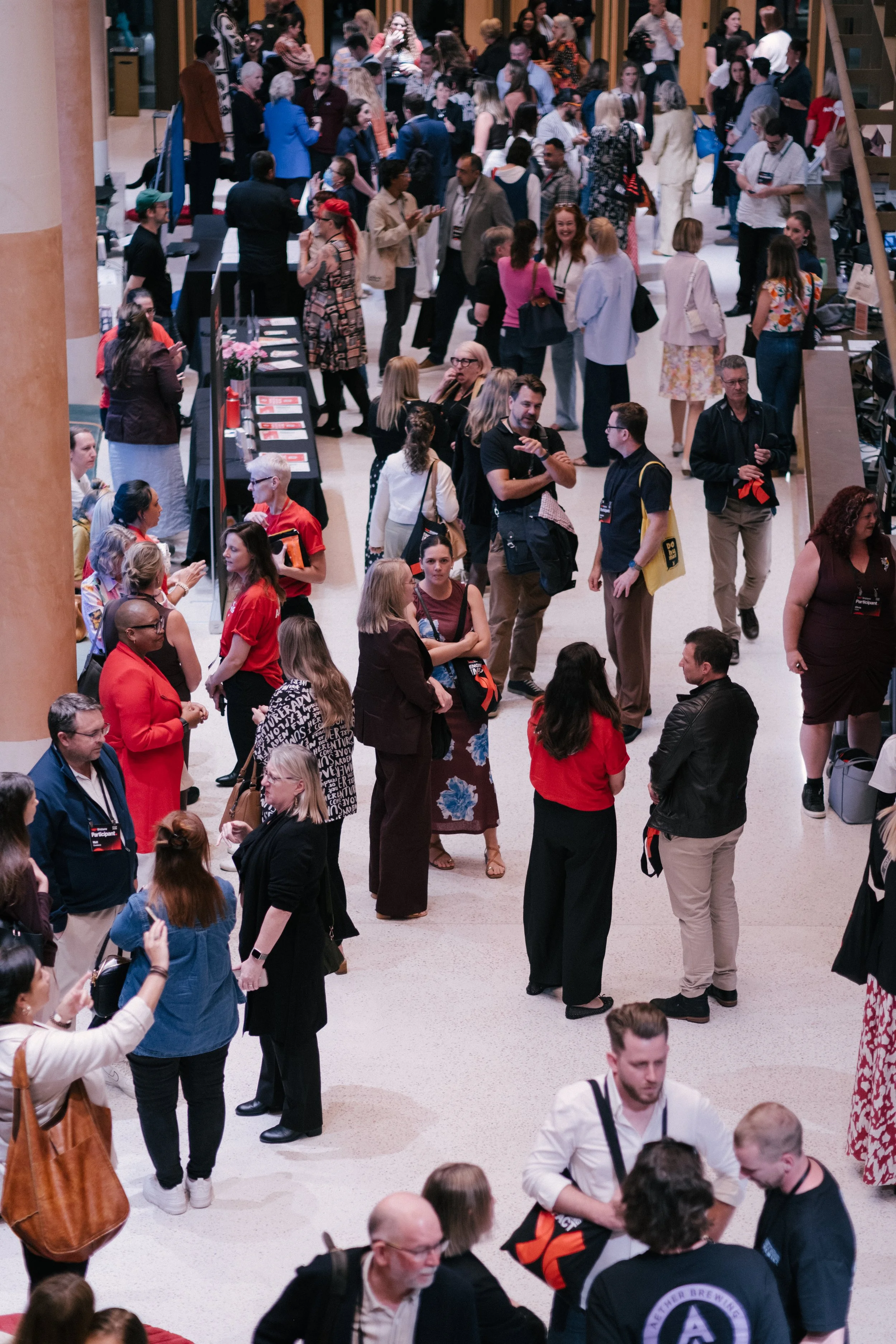 A crowded indoor event with many people standing around and talking, some looking at tables, in a large, well-lit room.
