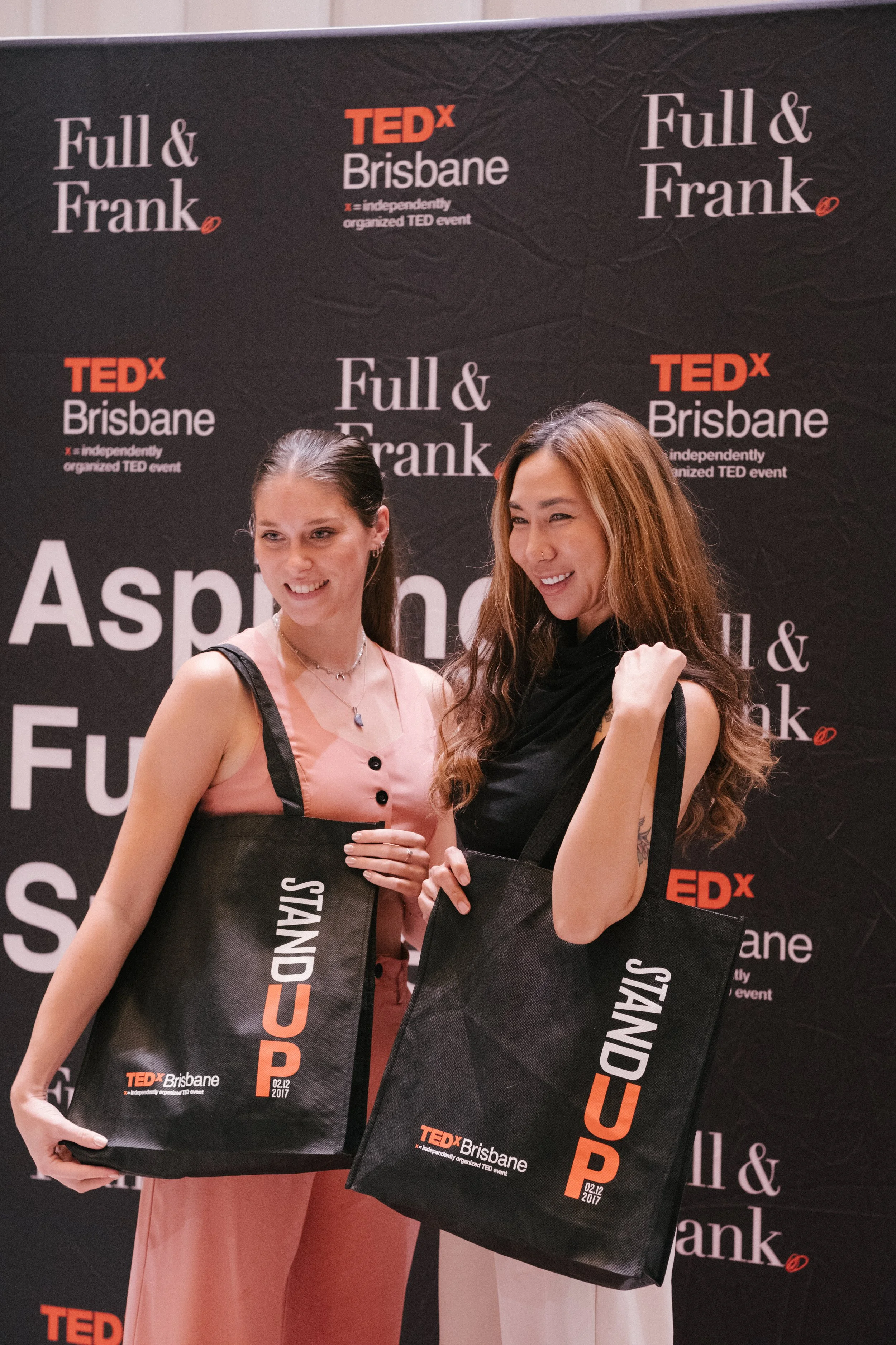 Two women at a TEDx Brisbane event, holding TEDx Brisbane tote bags, standing in front of a black backdrop with event logos.