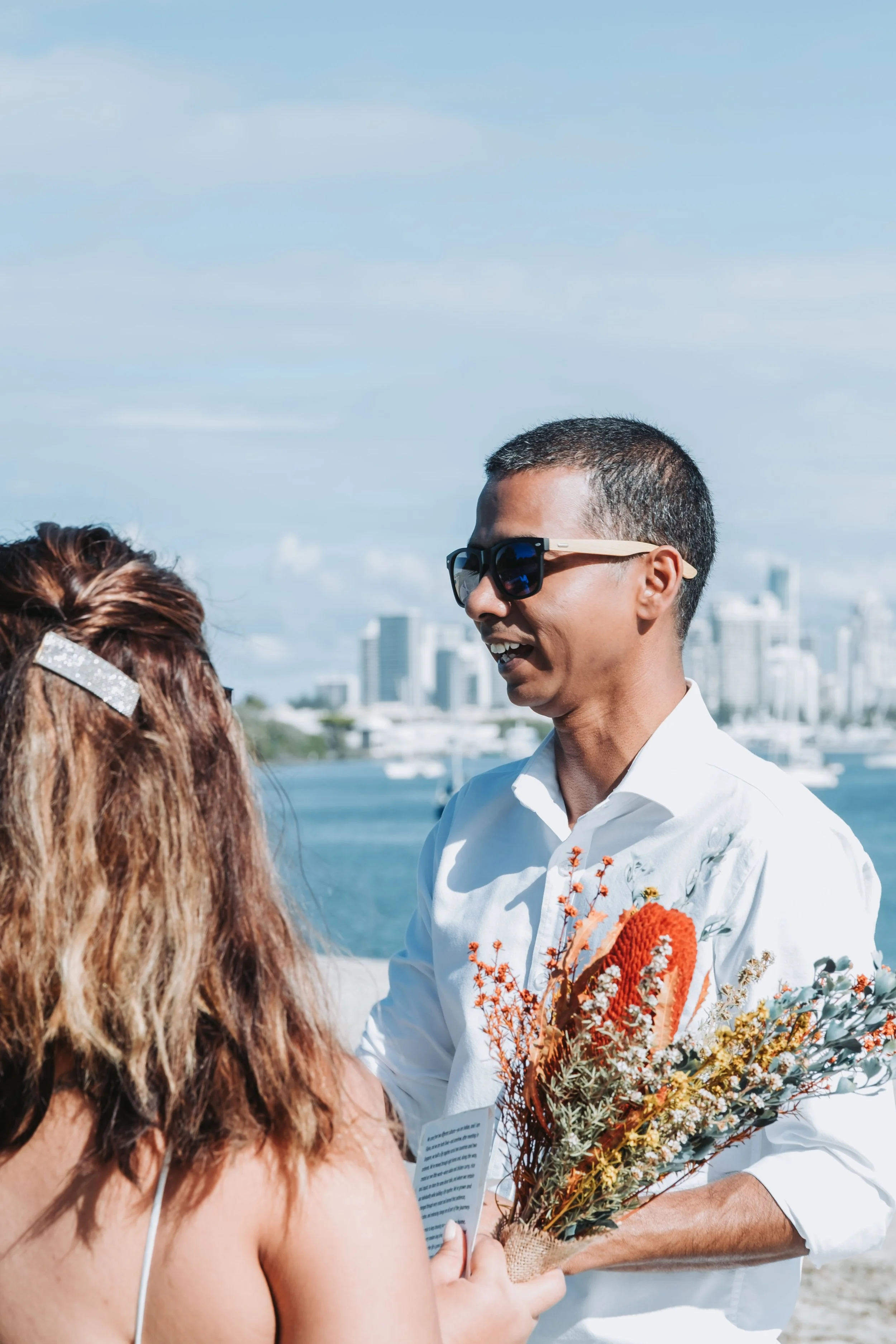 A man with short dark hair wearing sunglasses and a white shirt holding a bouquet of orange and white flowers during an outdoor event by the water with city buildings in the background.