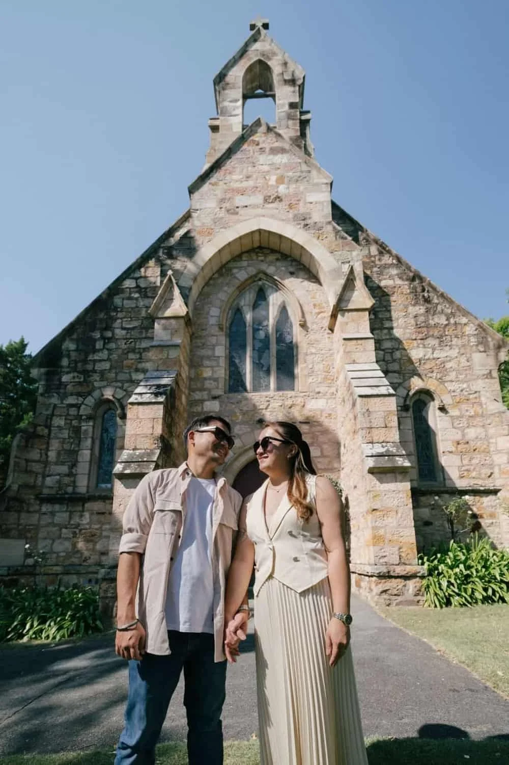 A couple holding hands and smiling in front of a stone church with Gothic-style windows and a steeple, on a sunny day.