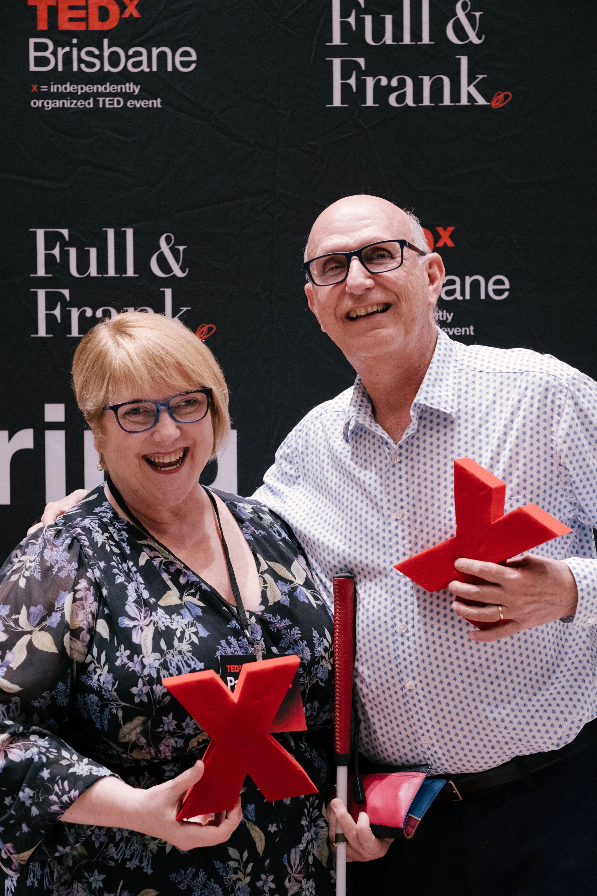 Two people, a woman and a man, smiling at a TEDx event in Brisbane. The woman has short blonde hair, glasses, and is wearing a floral dress. The man has glasses, a patterned shirt, and is holding a large red anvil-shaped trophy. They are standing in 