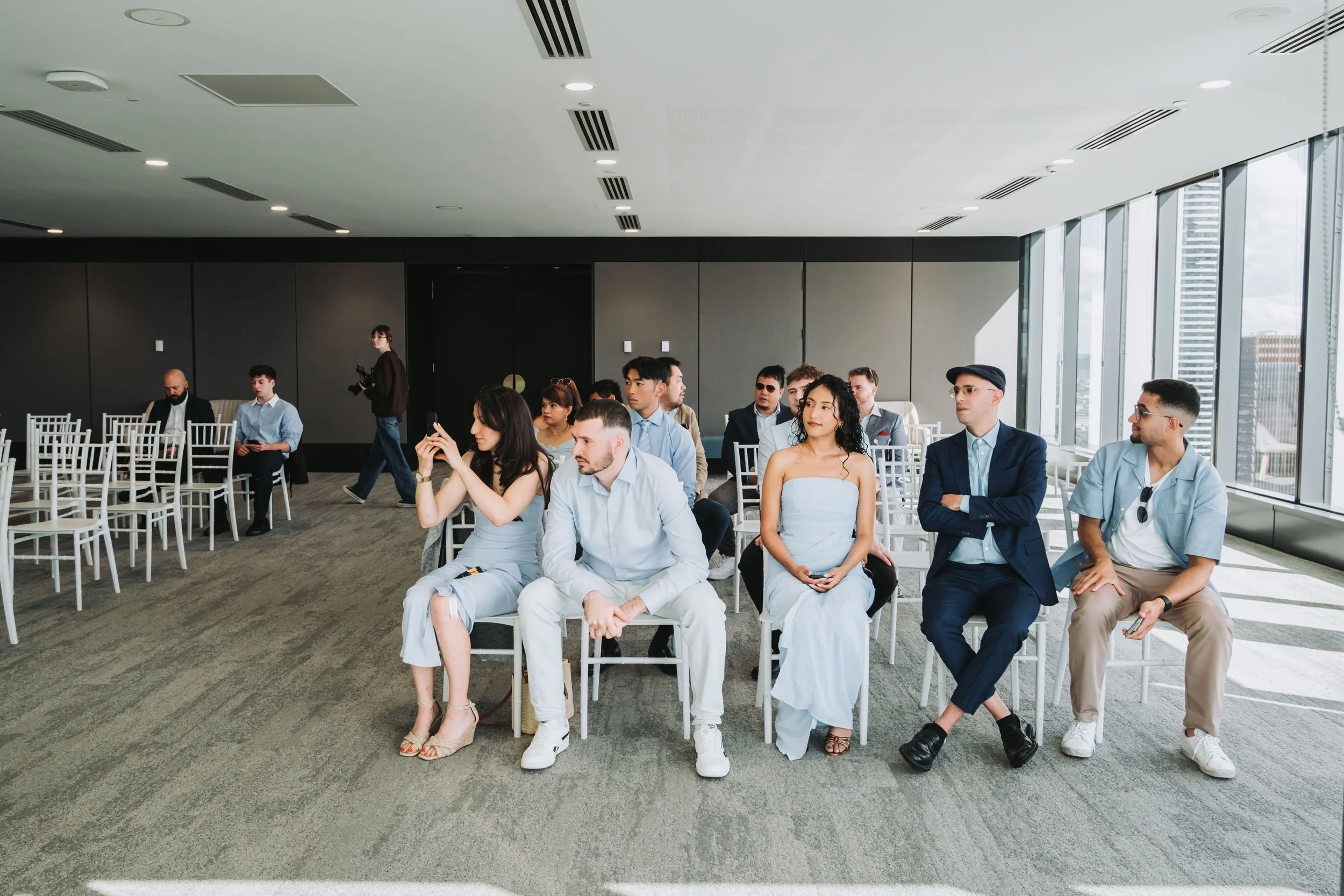 A group of diverse people sitting in a modern conference room with large windows, some using phones, others looking forward, and a woman standing with a camera, ready for an event or presentation.