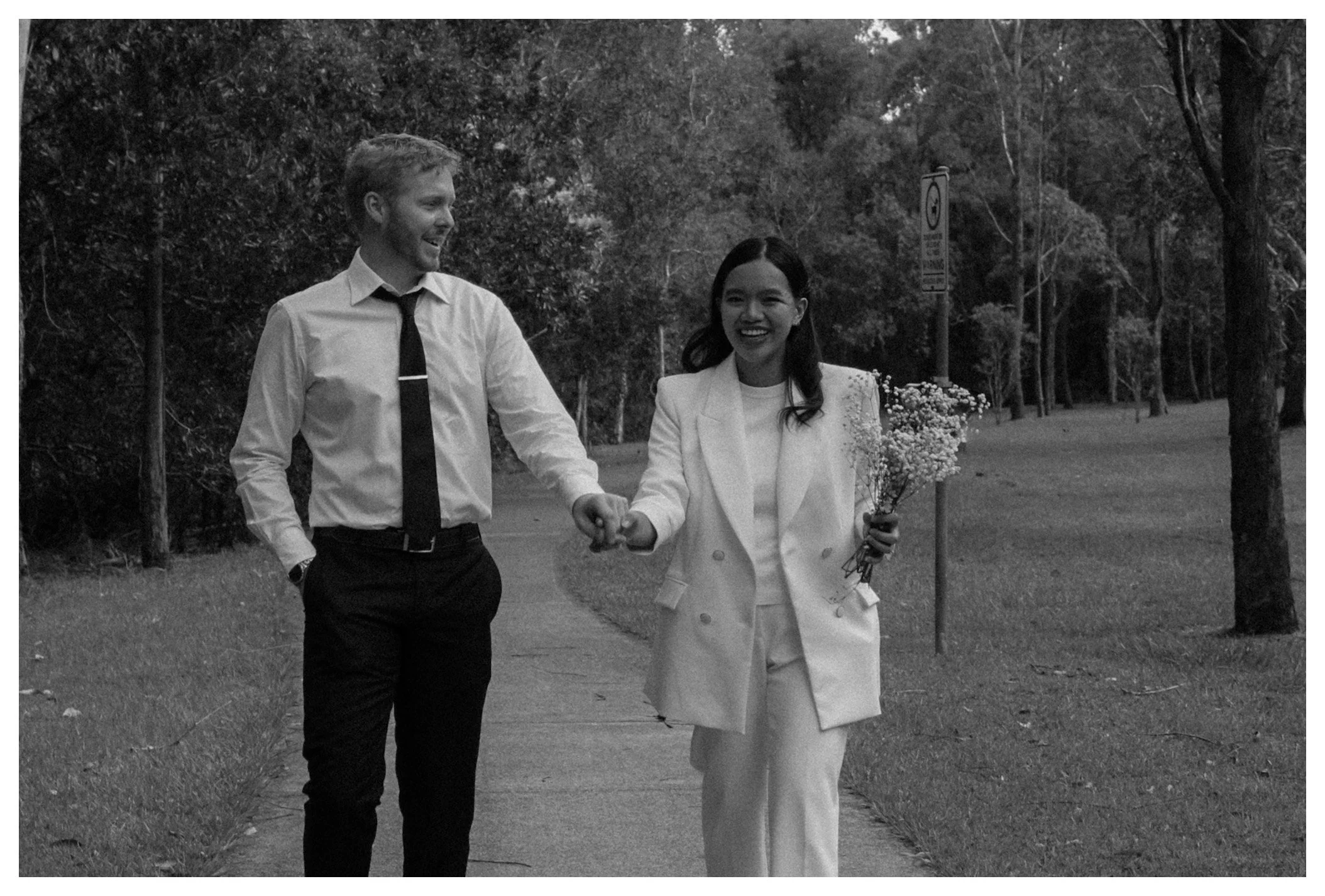 A man and a woman walking hand in hand outdoors on a park path, smiling and looking at each other. The woman is holding a bouquet of flowers. Trees surround them.