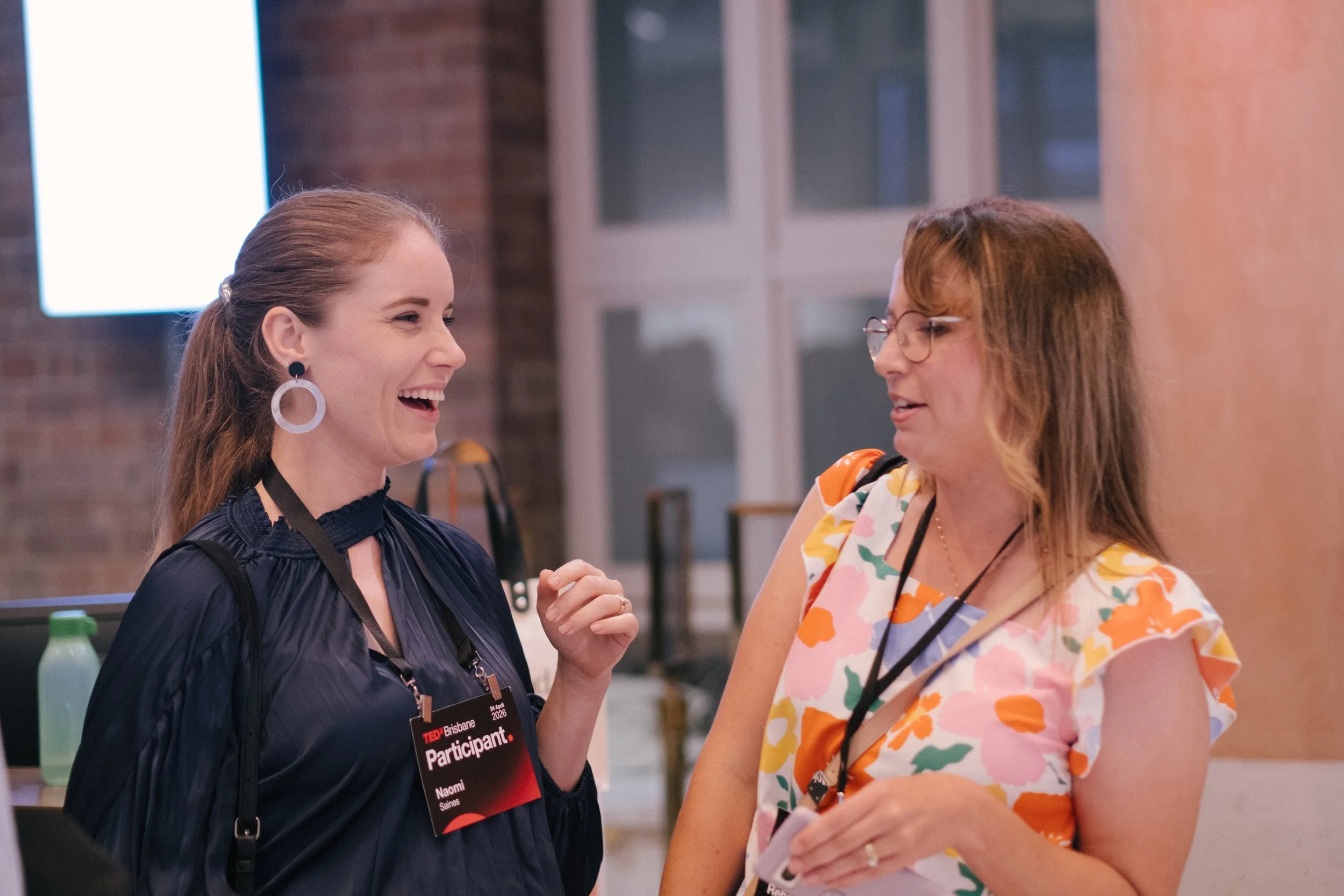 Two women are engaging in a conversation, smiling and holding hands at a busy event, with one wearing a black jacket and the other wearing a colorful patterned dress.
