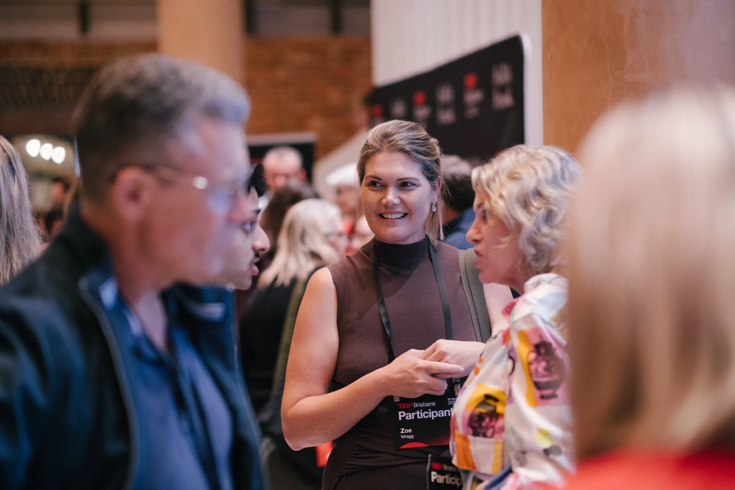 A woman with a 'Participant' badge engaging in conversation with an elderly woman at a TEDx event. The background shows a busy indoor setting with several other people.