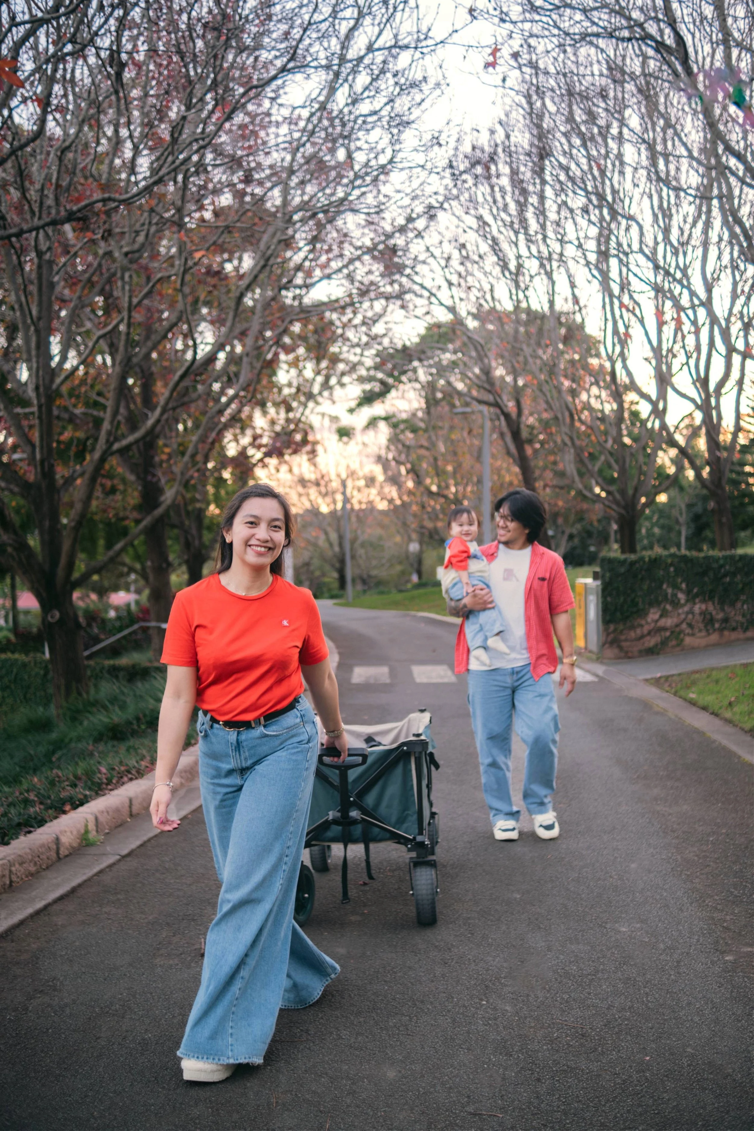 Three people walking in a park during sunset, two women and a young girl, one woman pushing a stroller.