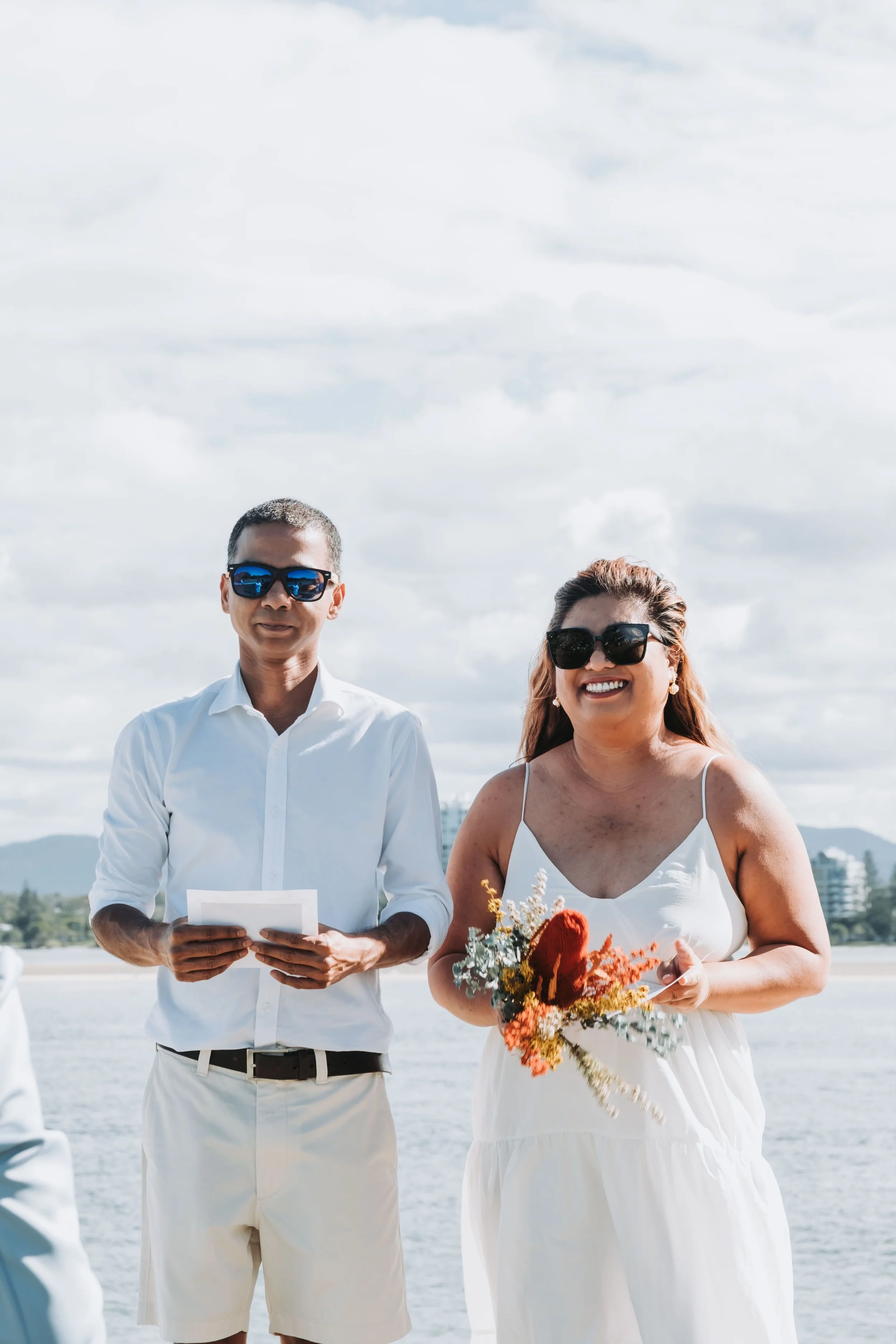 A man and woman in white clothing, both wearing sunglasses, standing outdoors on a cloudy day. The woman is holding a bouquet of flowers, and the man is holding a piece of paper, possibly during a wedding or similar event.