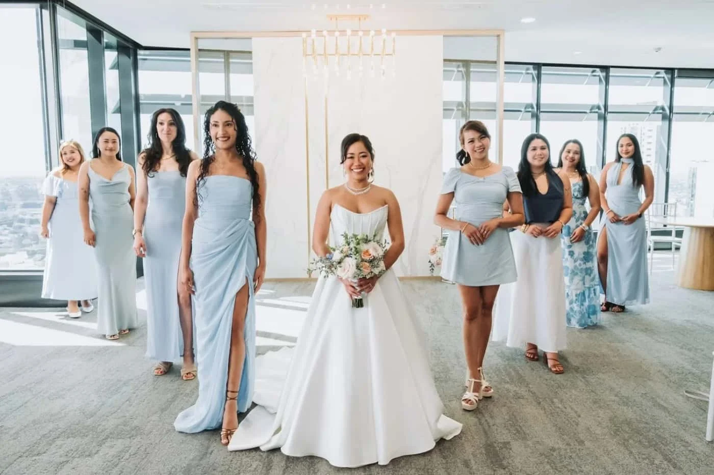 A bride in a white wedding dress holding a bouquet surrounded by bridesmaids in light blue and pastel dresses in a modern high-rise building with large windows and city views.