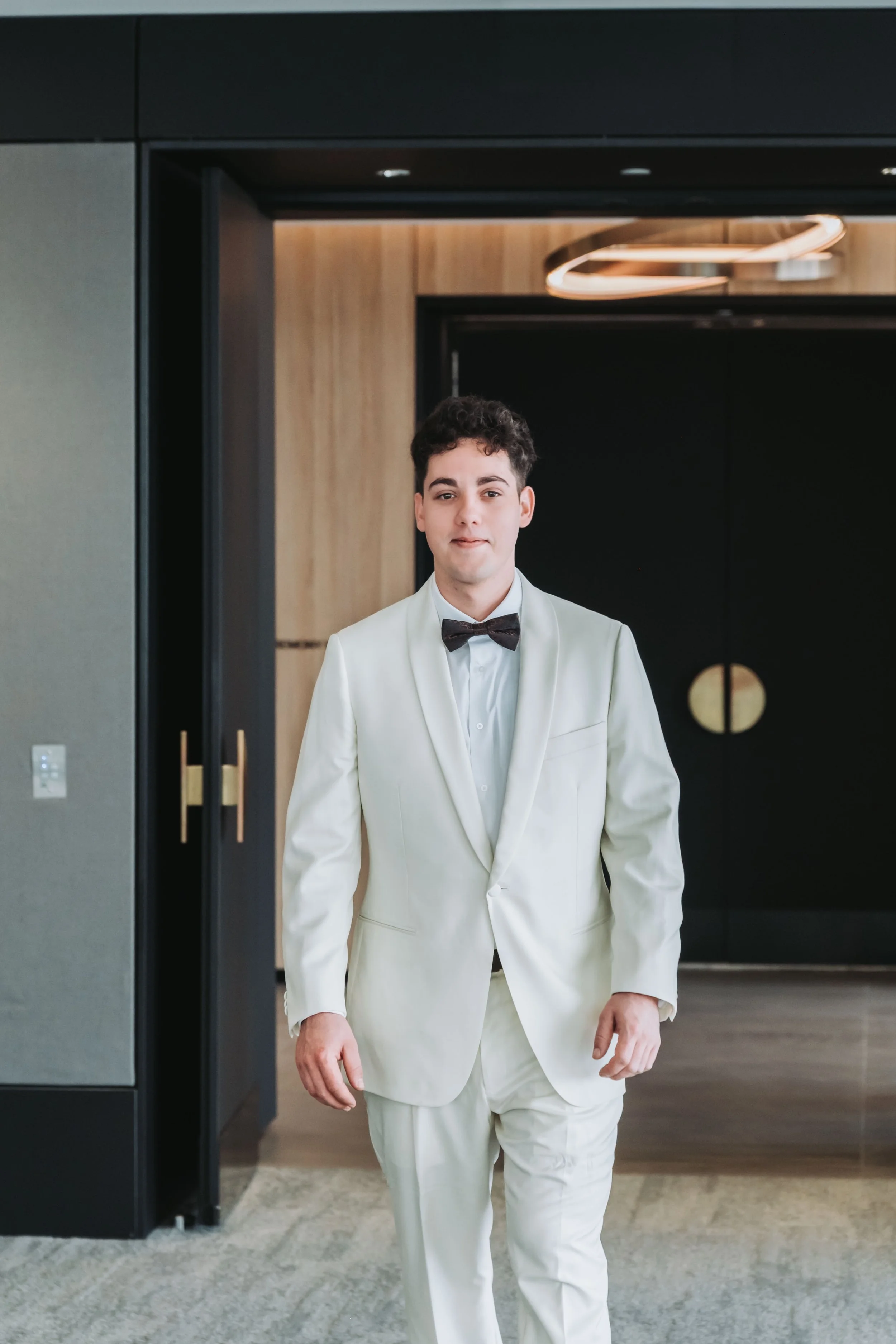 A young man in a white suit and black bow tie standing in front of a modern black and wood background.