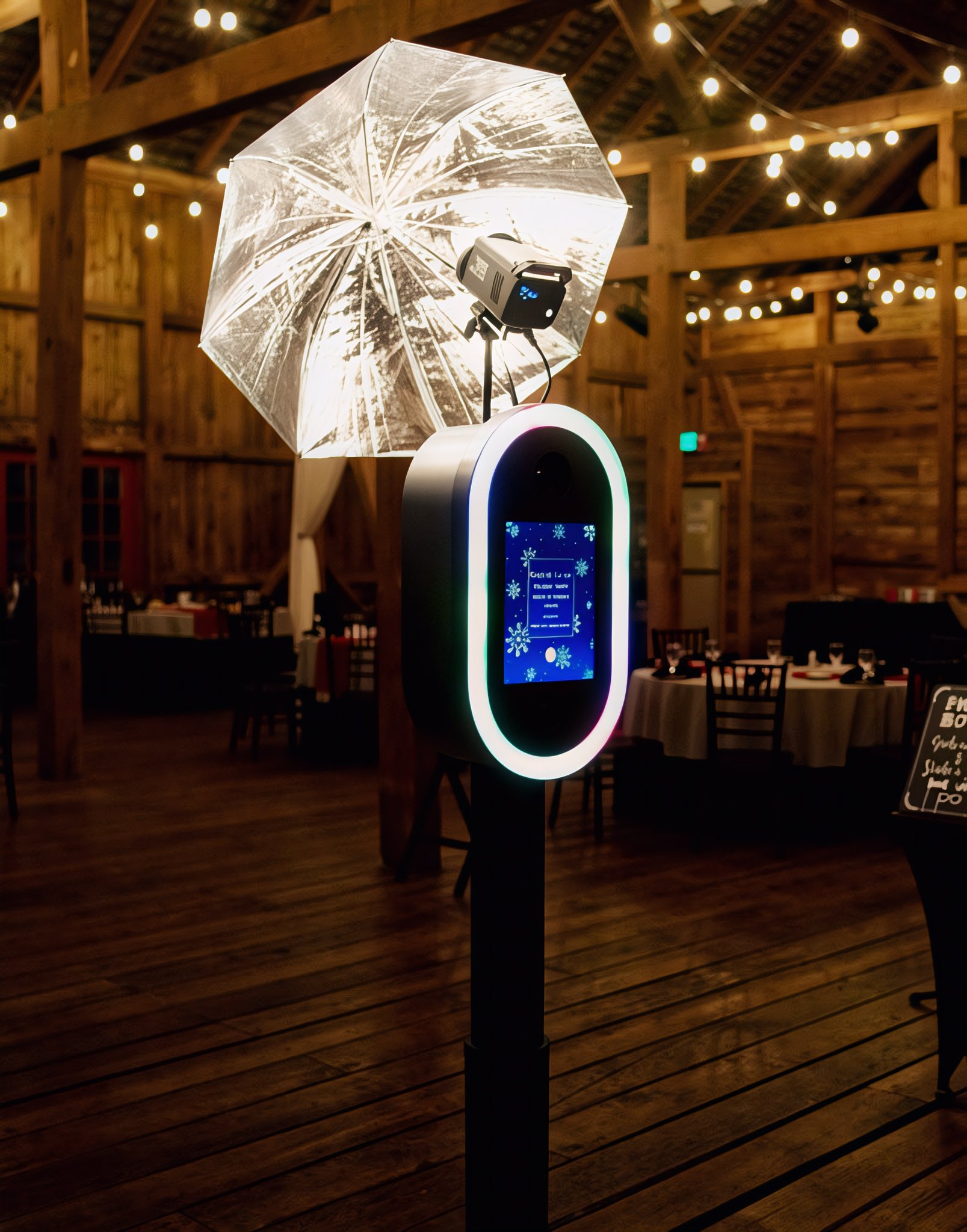 Indoor event space with wooden beams and string lights, featuring a photo booth with a digital display and a snowflake decoration, and a stand-up umbrella with an LED light.
