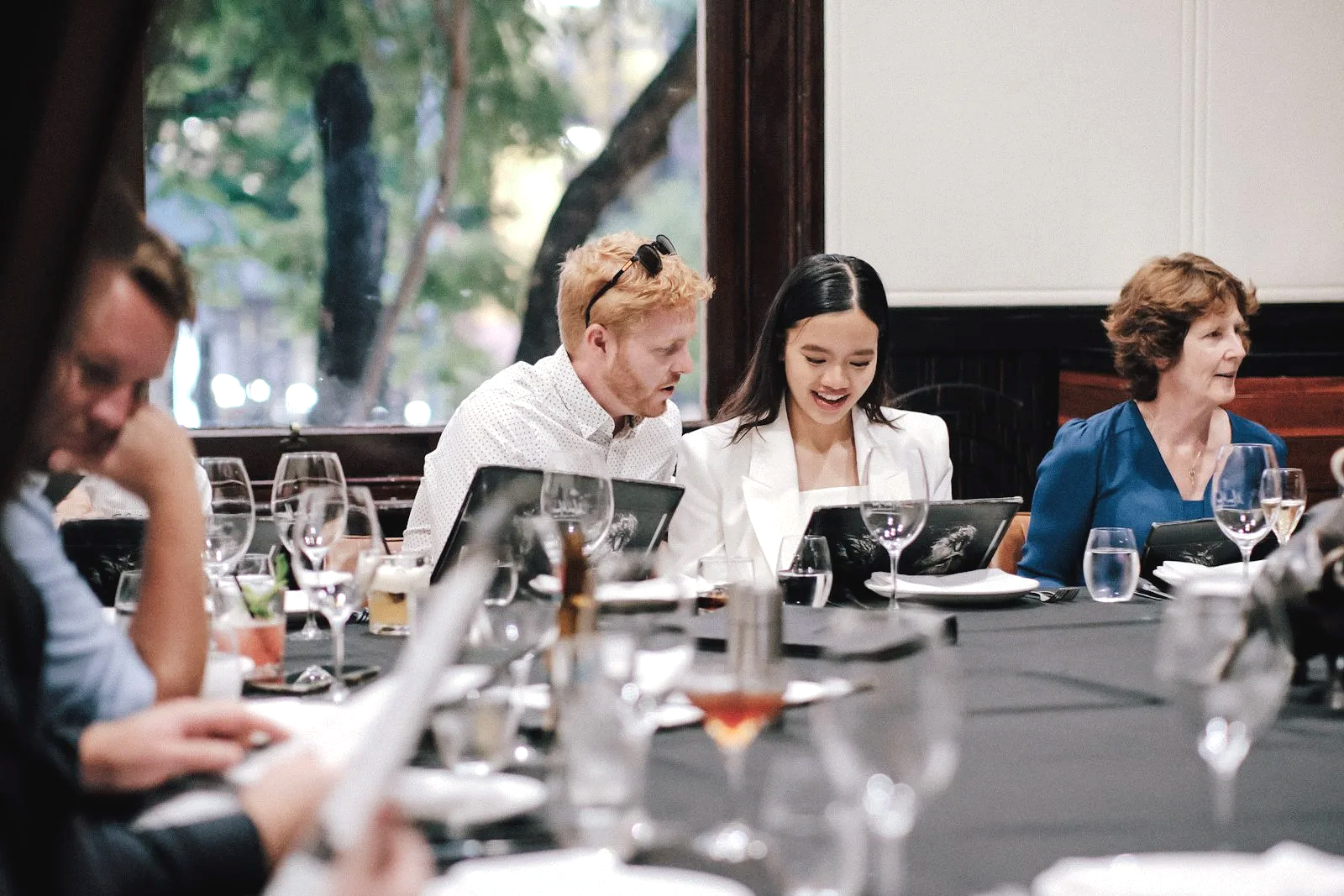 A group of people sitting at a table in a restaurant, dressed in business or formal attire, looking at menus and enjoying a meal. The background shows a window with trees outside.