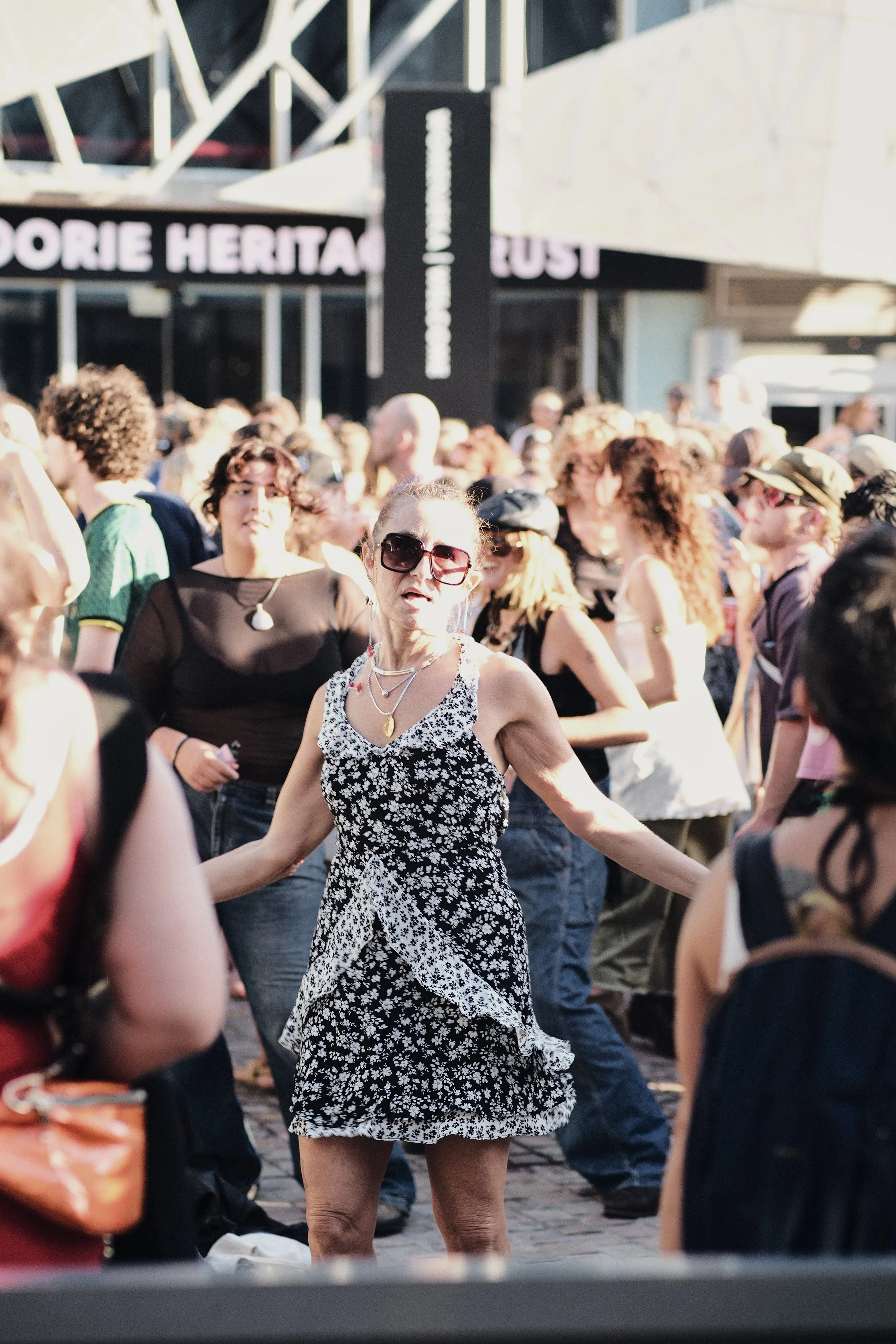A woman with sunglasses and a floral dress dancing or celebrating in a crowd at an outdoor event.