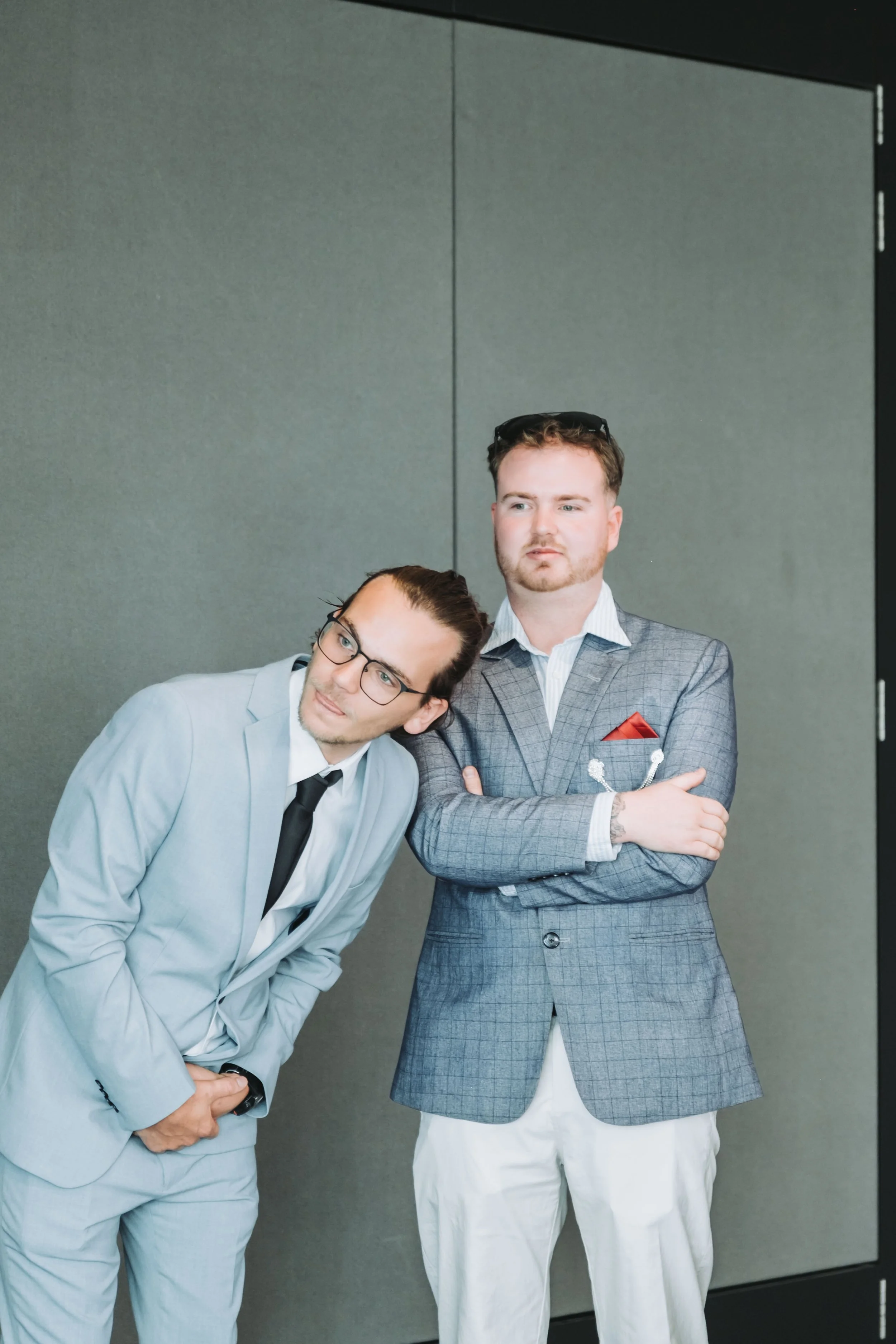 Two men in suits stand together against a plain wall. One man, on the left, is leaning his head on the shoulder of the other man, on the right. Both are dressed in light-colored suits, with the man on the right wearing a gray plaid suit with a red po
