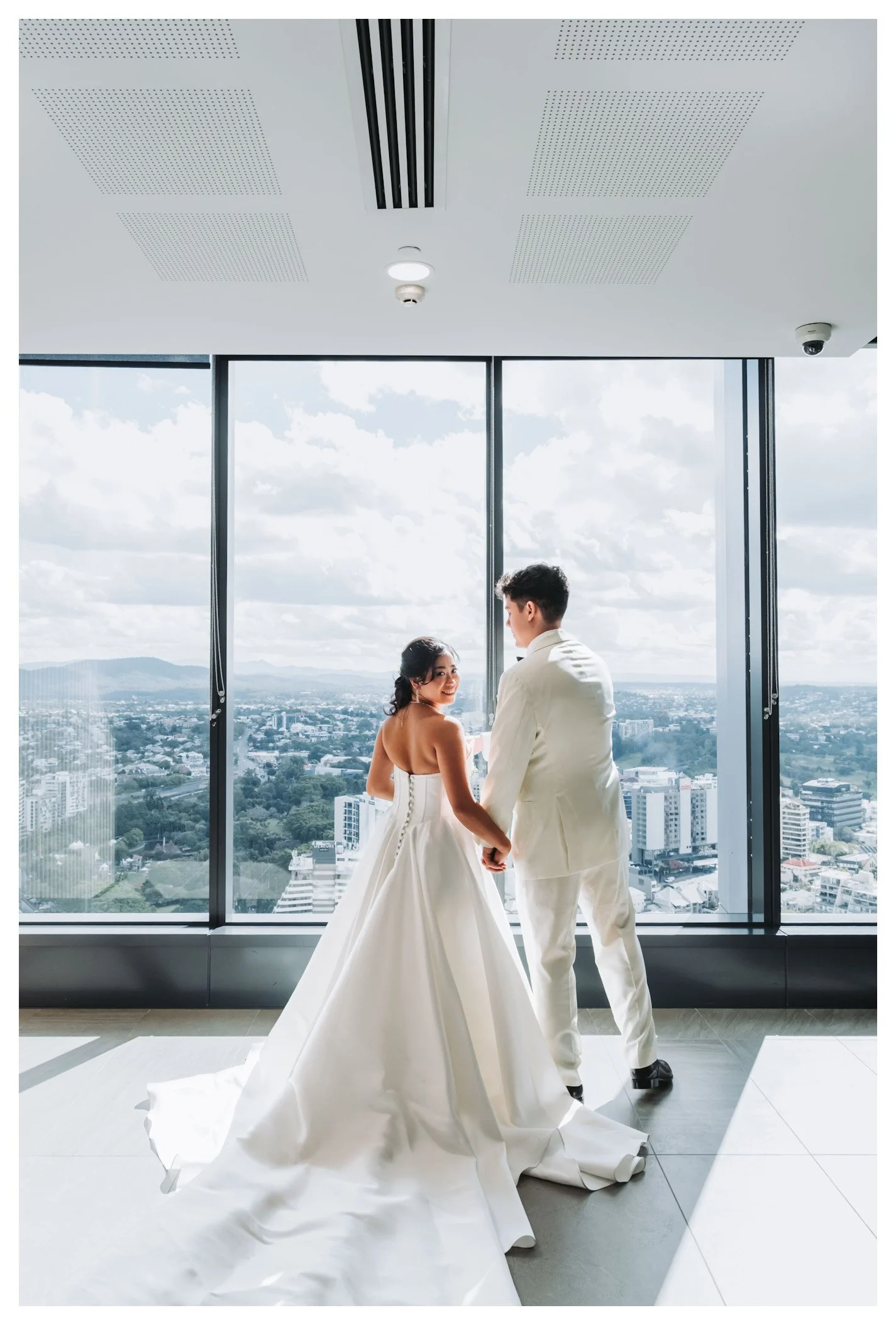 Bride and groom holding hands in white wedding attire, standing before floor-to-ceiling windows with city skyline view.