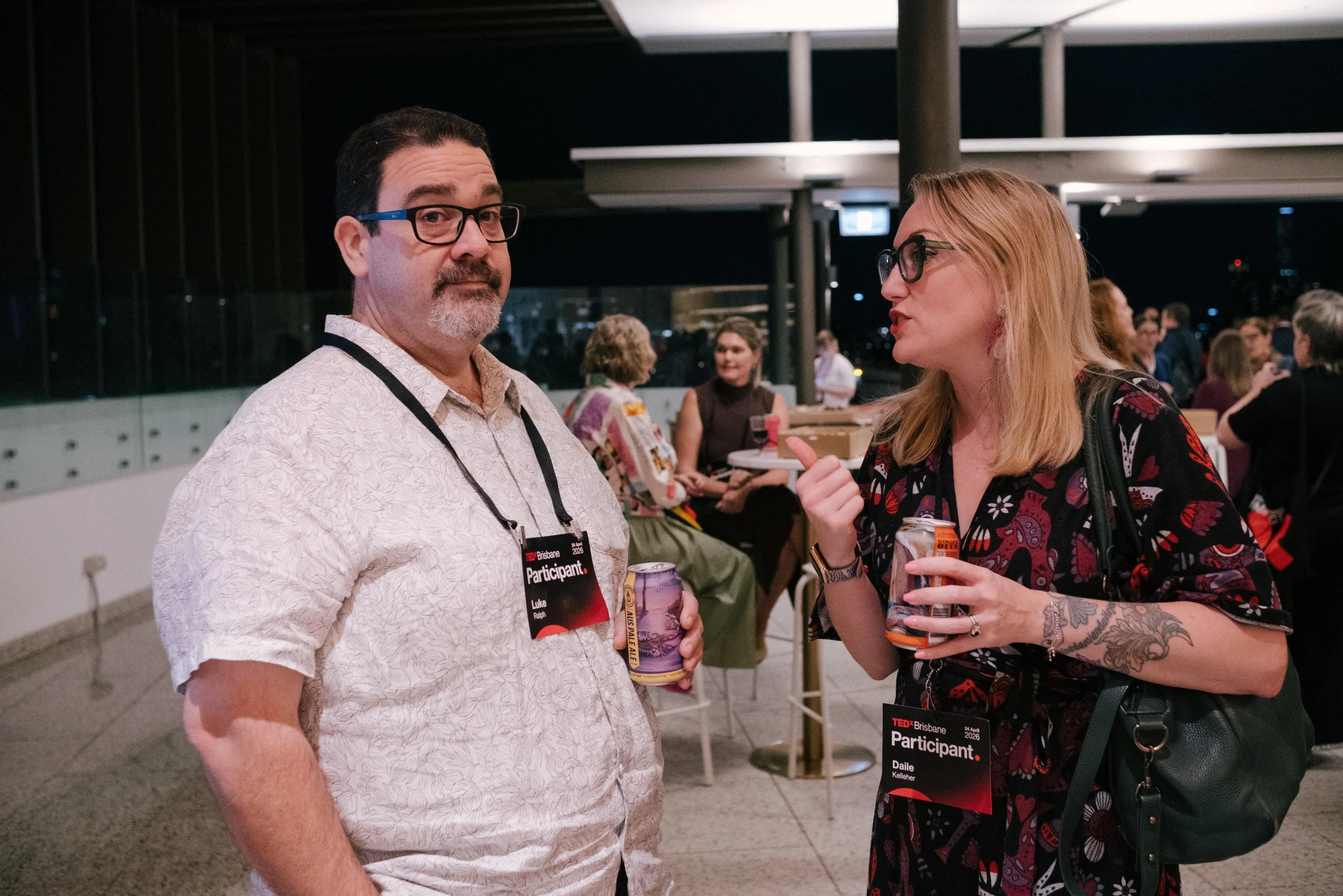 Two people at a TEDx Brisbane event socializing indoors at night, both wearing event nametags and holding drinks, with more attendees in the background.