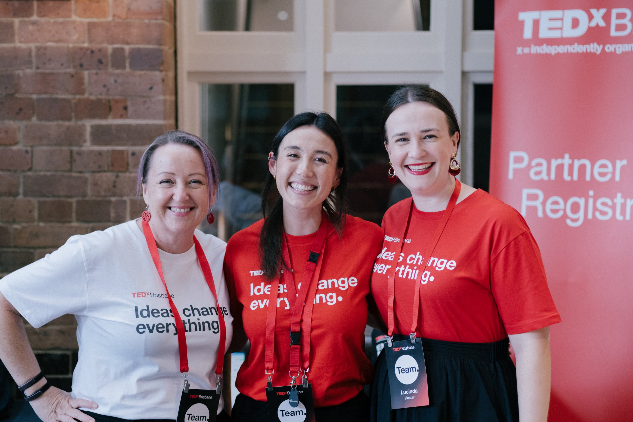 Three women standing together at a TEDx event, smiling. Two women wear red shirts with white text, and one wears a white shirt with black text. They have red lanyards with badges that say "Team". There is a pink TEDx banner in the background and a brick wall on the left.