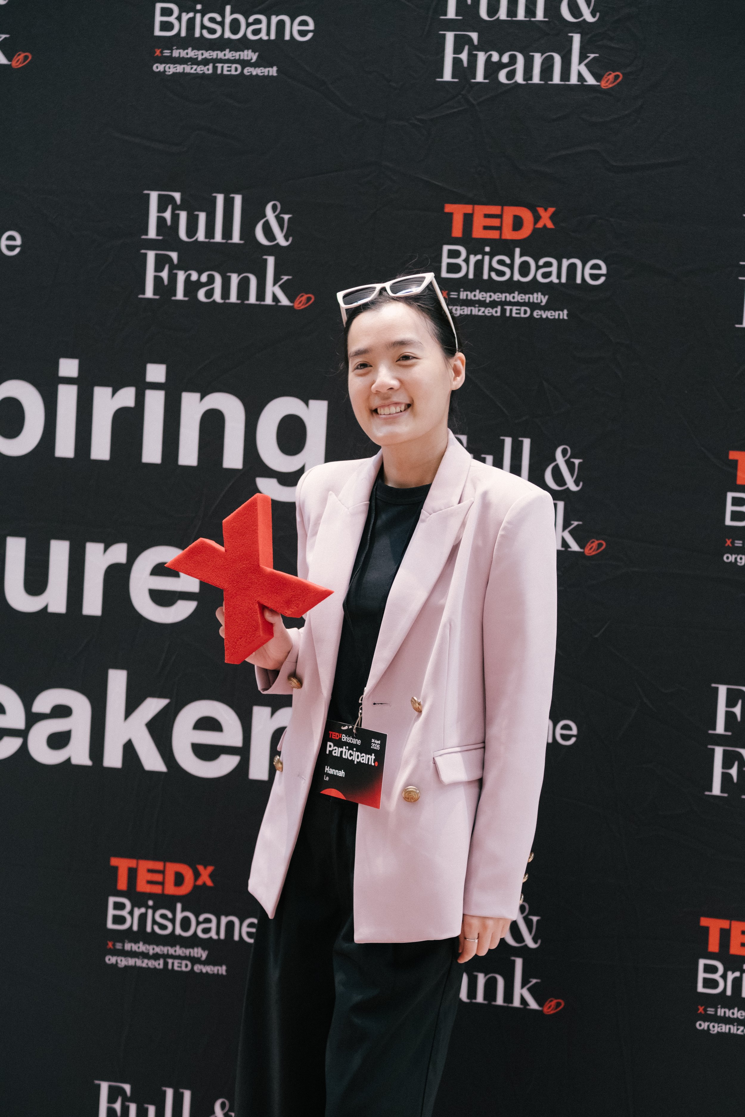 A woman standing in front of a black TEDx Brisbane backdrop, smiling, holding a red star-shaped award, wearing glasses on her head, a light pink blazer, black pants, and a badge that says 'Participant' with her name, Hannah.