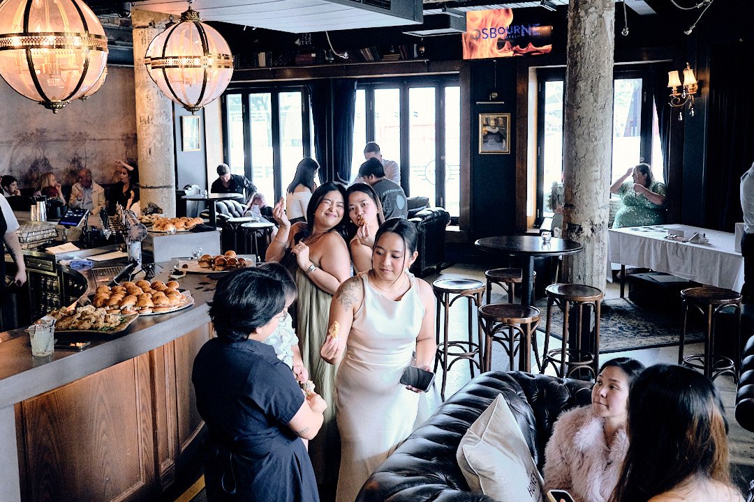 Women socializing at a café with food and drinks, with some seated at tables and others chatting near the bar area.