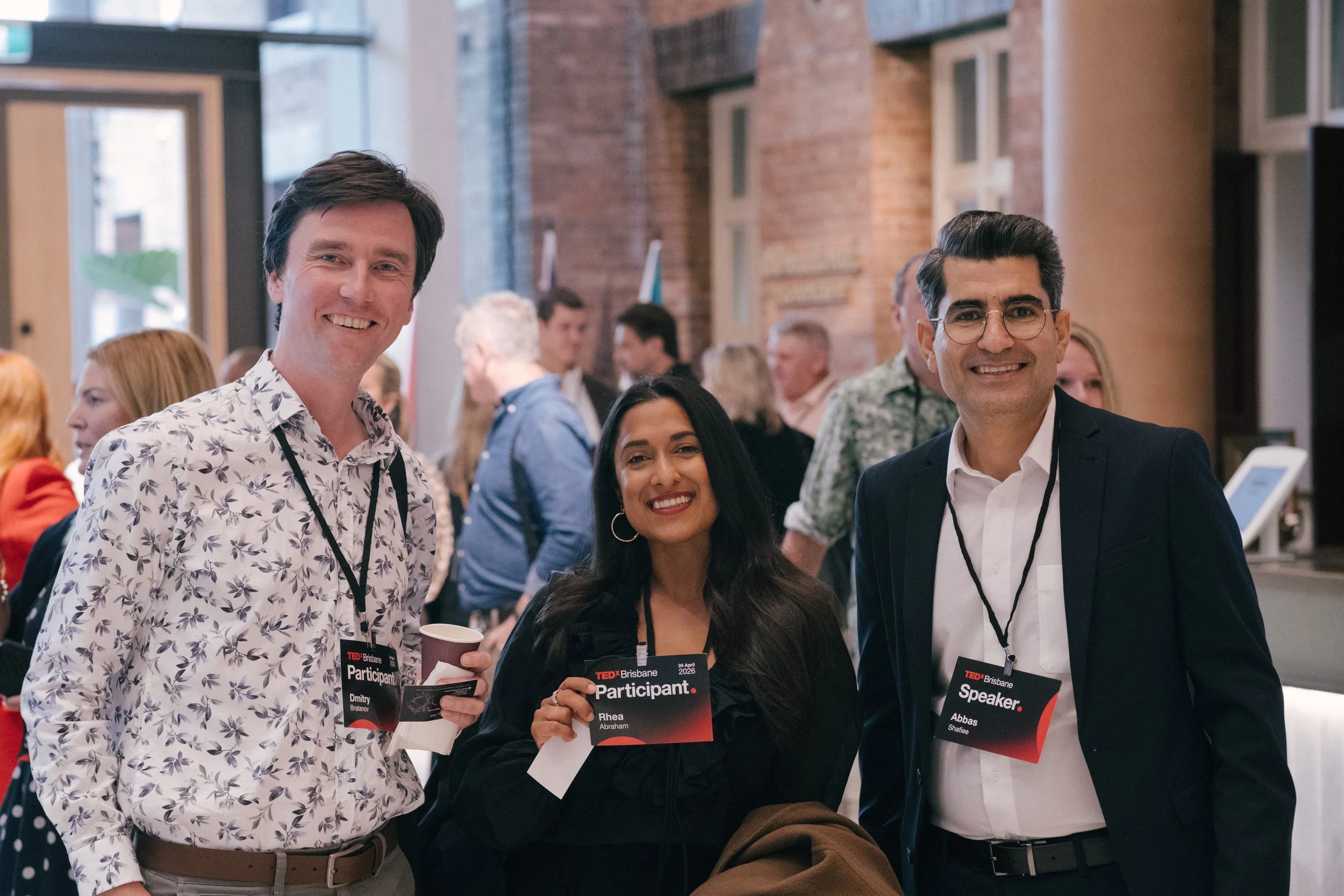 Three people smiling at an indoor event, holding TEDxBrisbane Participant and Speaker badges, with many attendees in the background inside a brick-walled room.
