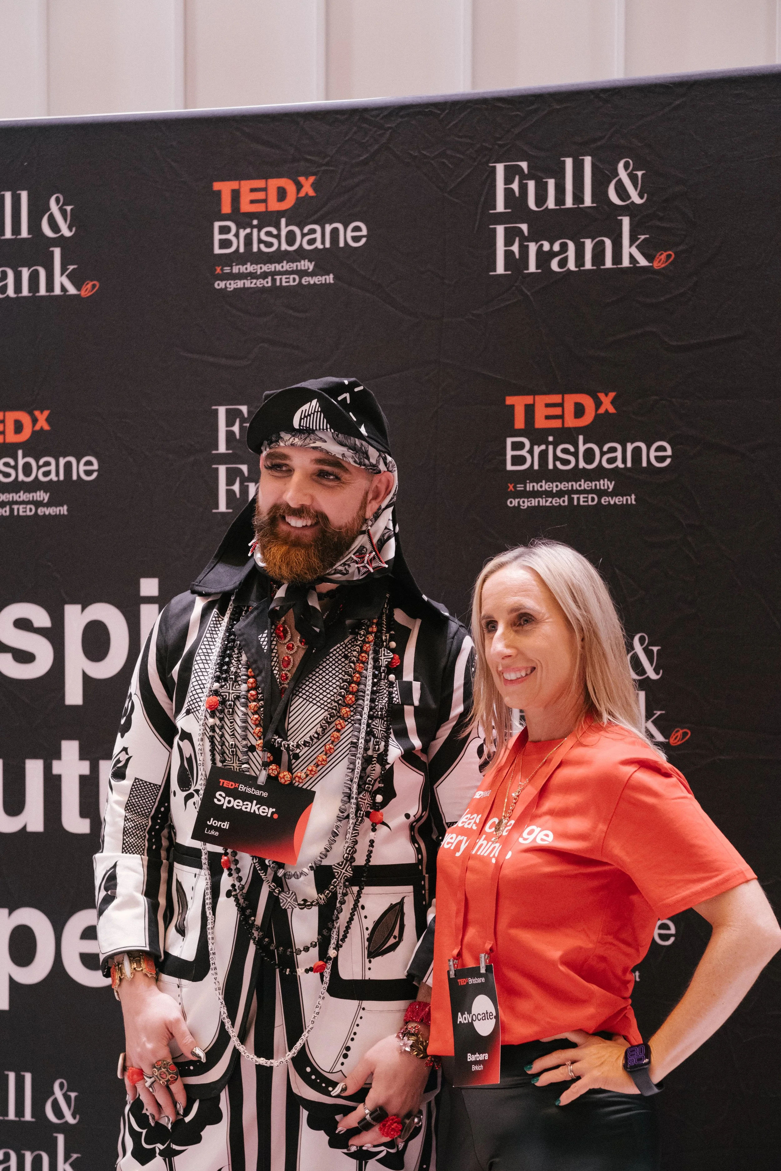 Two women standing together at a TEDx Brisbane event, smiling. The woman on the left is wearing a patterned black and white outfit with numerous necklaces, rings, and bracelets, and has a name tag that reads 'Jordi Luke'. She has a beard and is dress