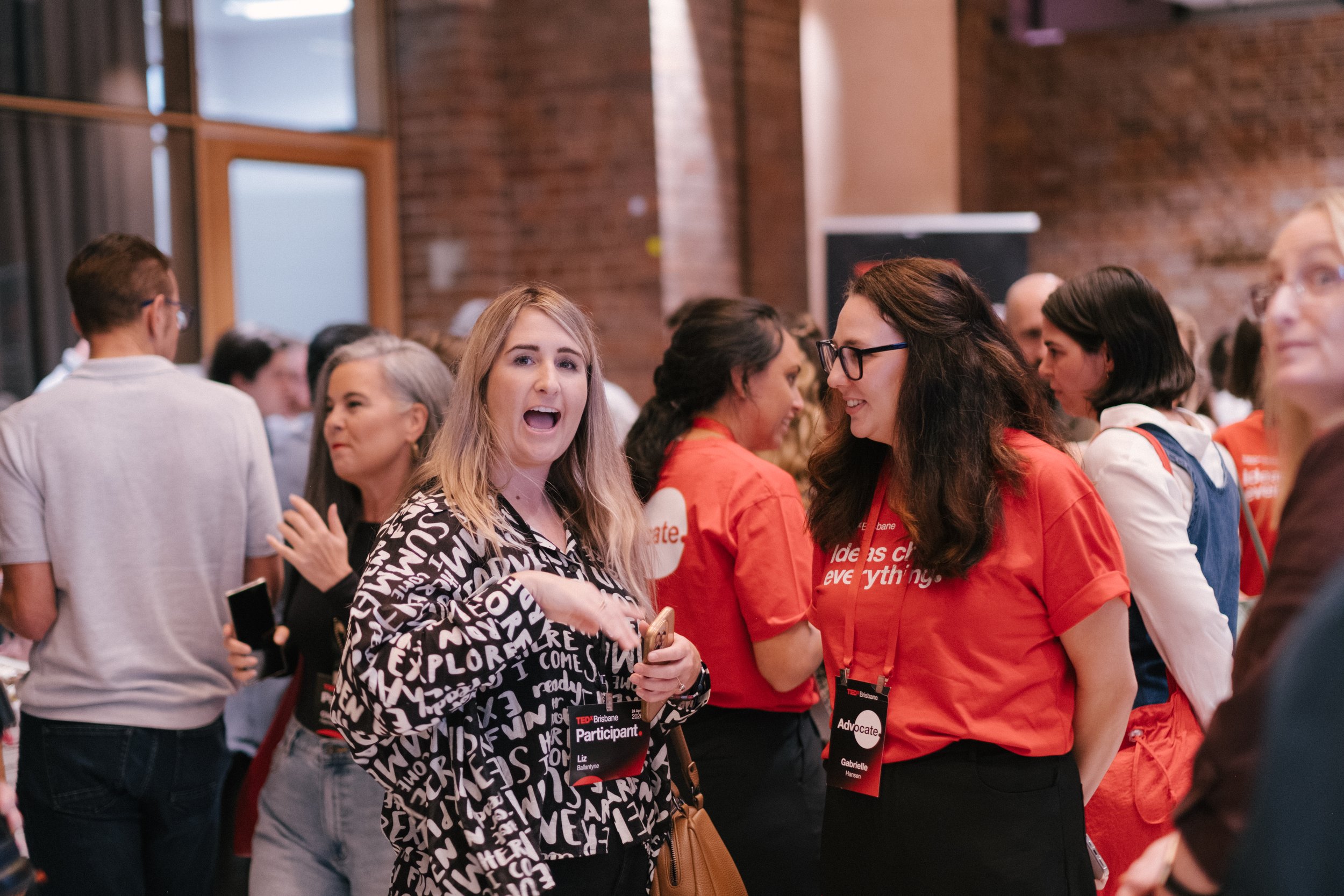 A group of people at a conference or event, with some individuals wearing name tags. Two women in the foreground are engaged in conversation, one with glasses and dark hair, the other with blonde hair and a black-and-white patterned top. The background shows a brick wall and other attendees.