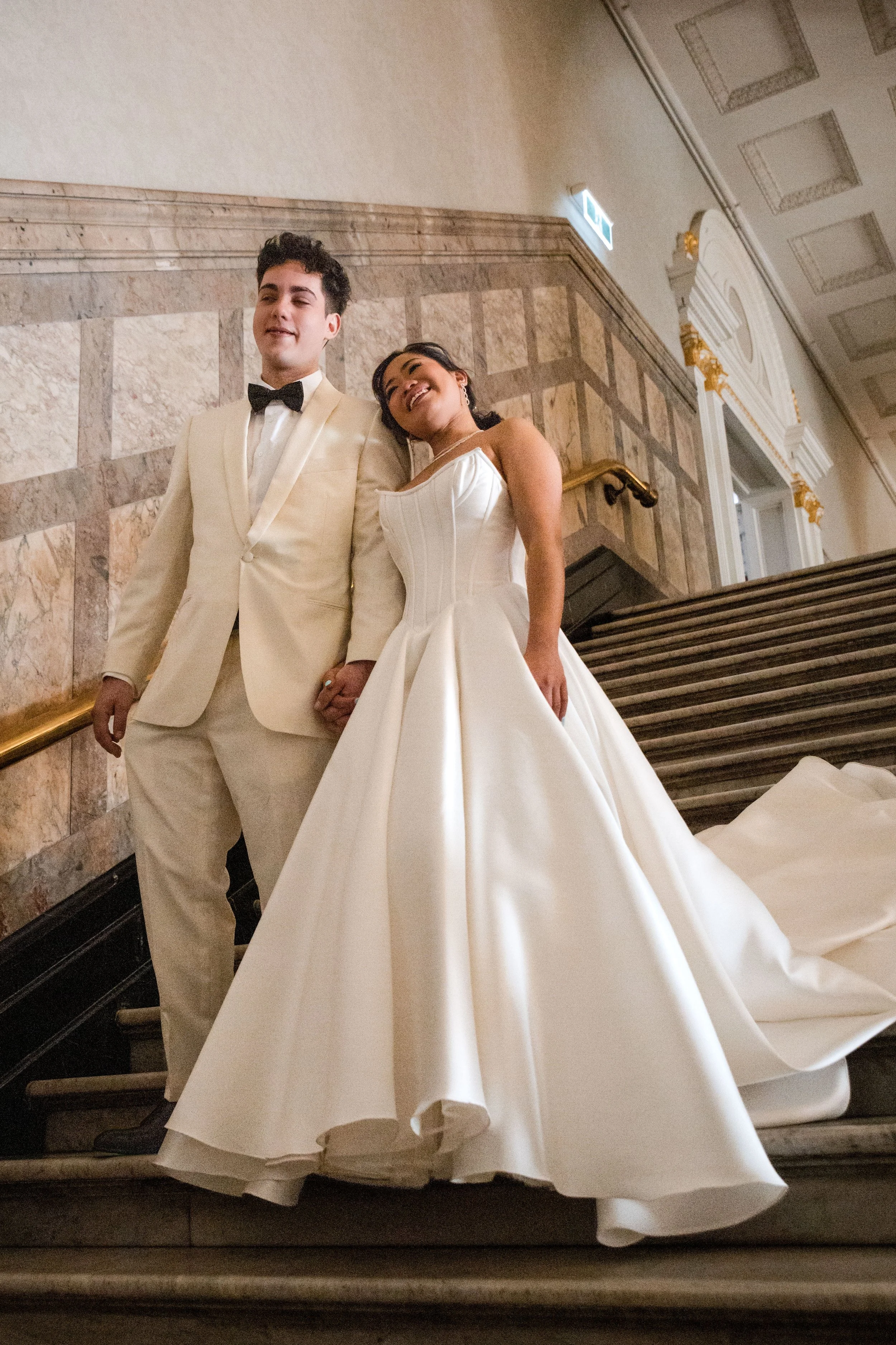 A happy couple dressed in formal wedding attire, holding hands and smiling, standing on a staircase inside a grand building with marble walls and ornate decor.