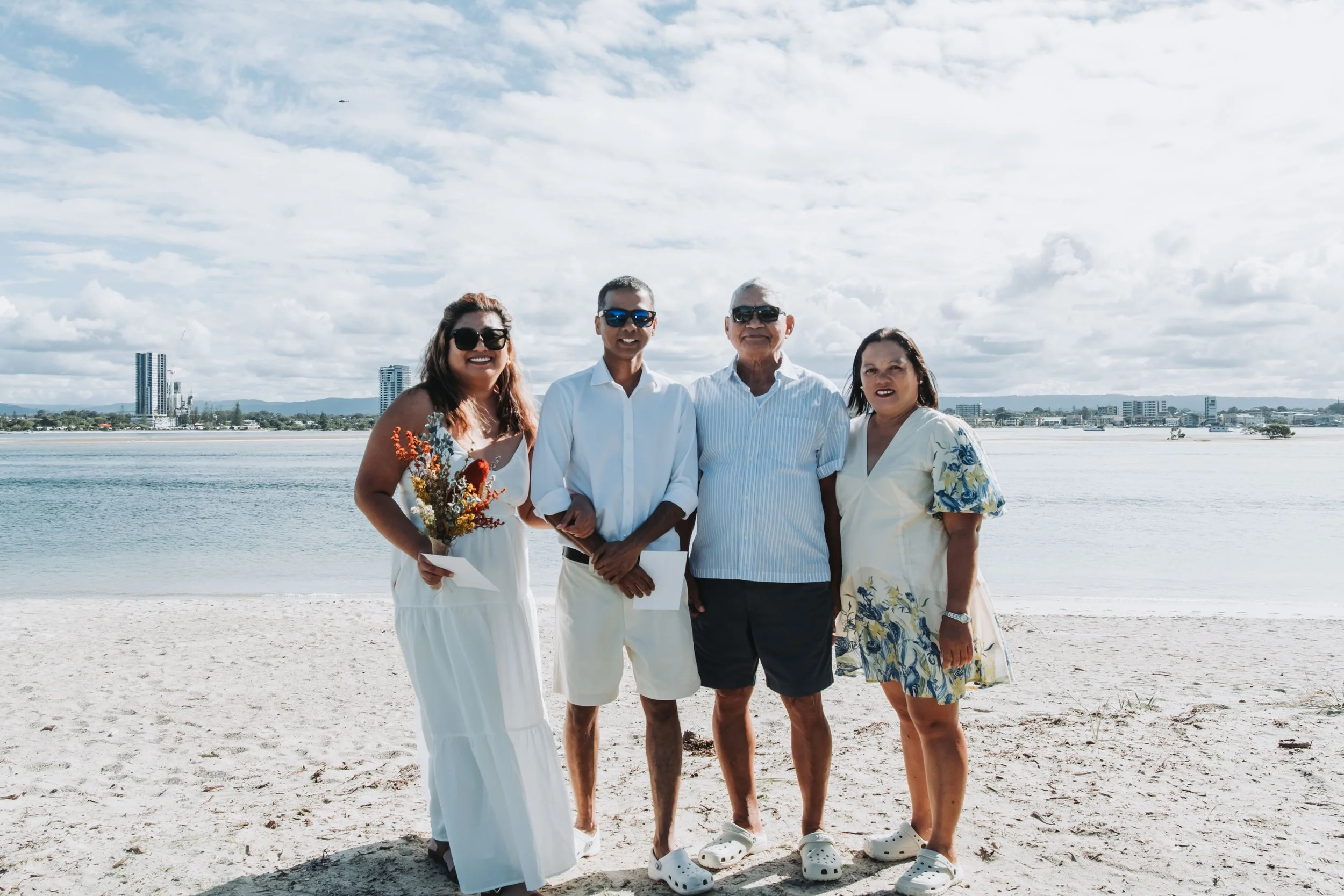 Four people standing on a sandy beach with buildings and water in the background, smiling at the camera, dressed in white and light-colored clothing.