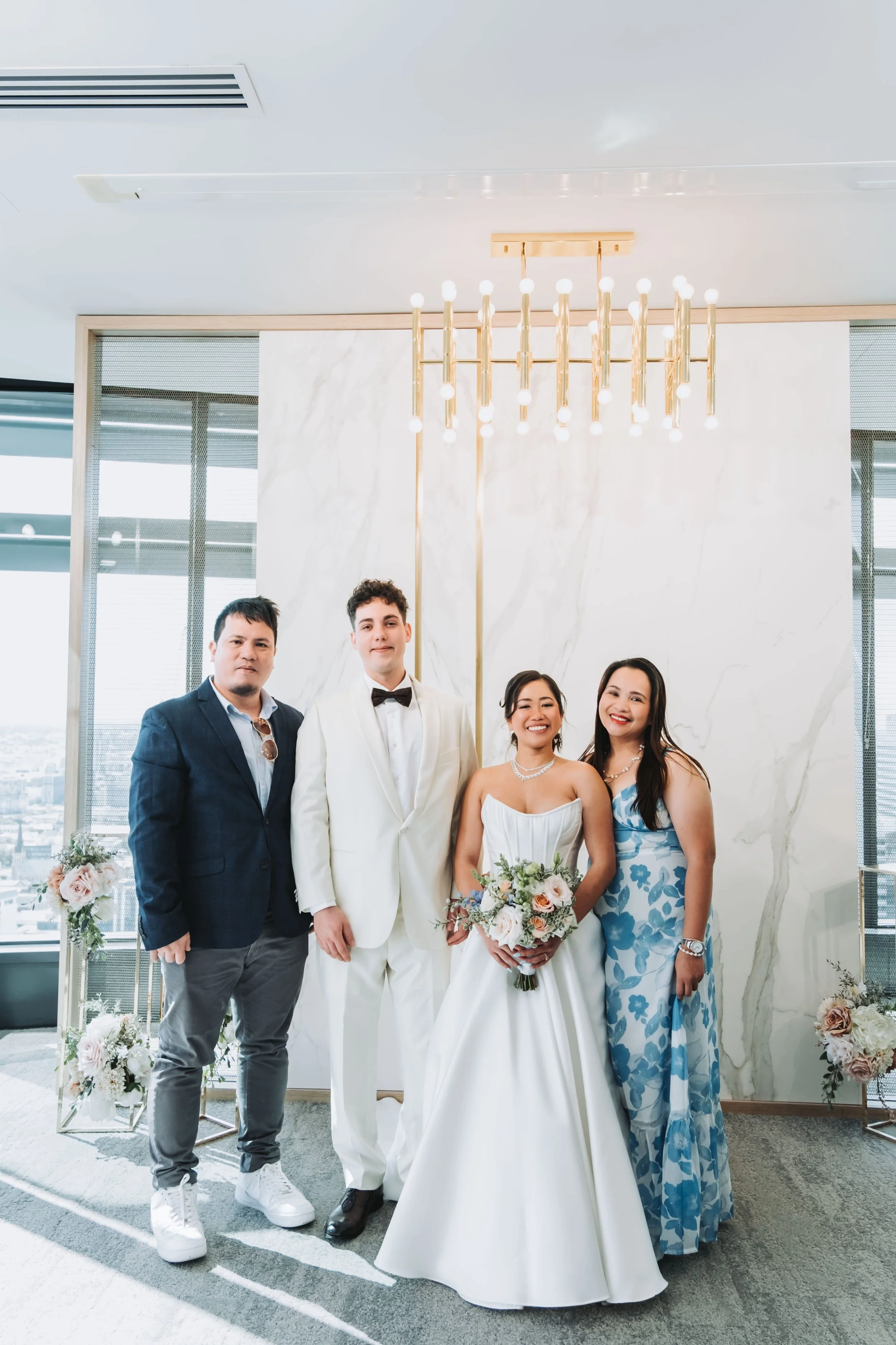 Group of five people, including a bride in a white wedding dress and veil holding a bouquet, and four others in formal attire, standing together in a bright, modern interior with large windows and contemporary light fixture, possibly at a wedding or 