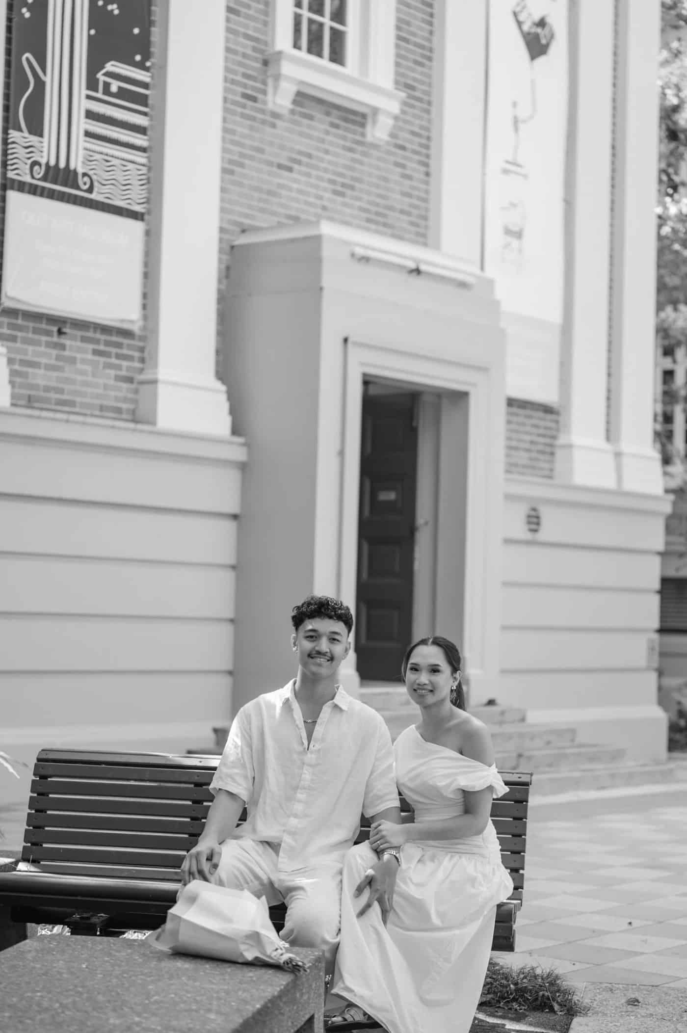 A young man and woman sitting on a park bench in front of a house, smiling at the camera. The man is wearing a light-colored shirt and light pants, and the woman is dressed in a white dress. There is a paper bag on the ground near their feet.