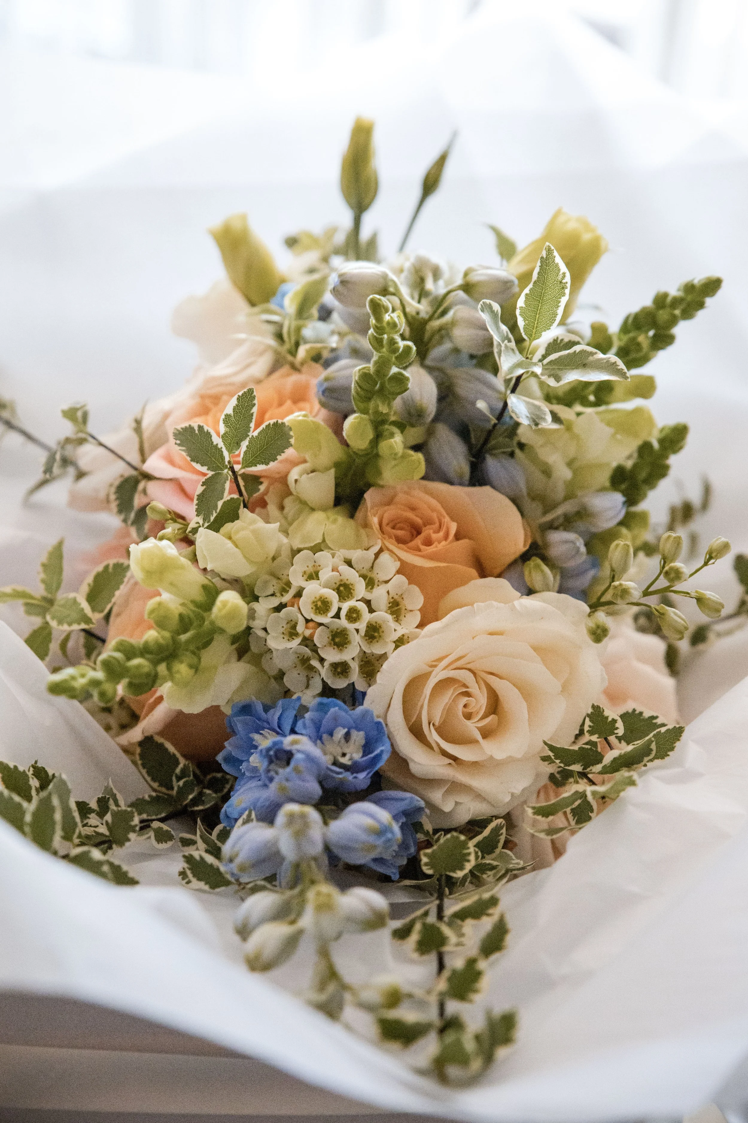 A bouquet of mixed flowers including ivory roses, small white blossoms, blue flowers, and greenery with variegated leaves, wrapped in white paper.