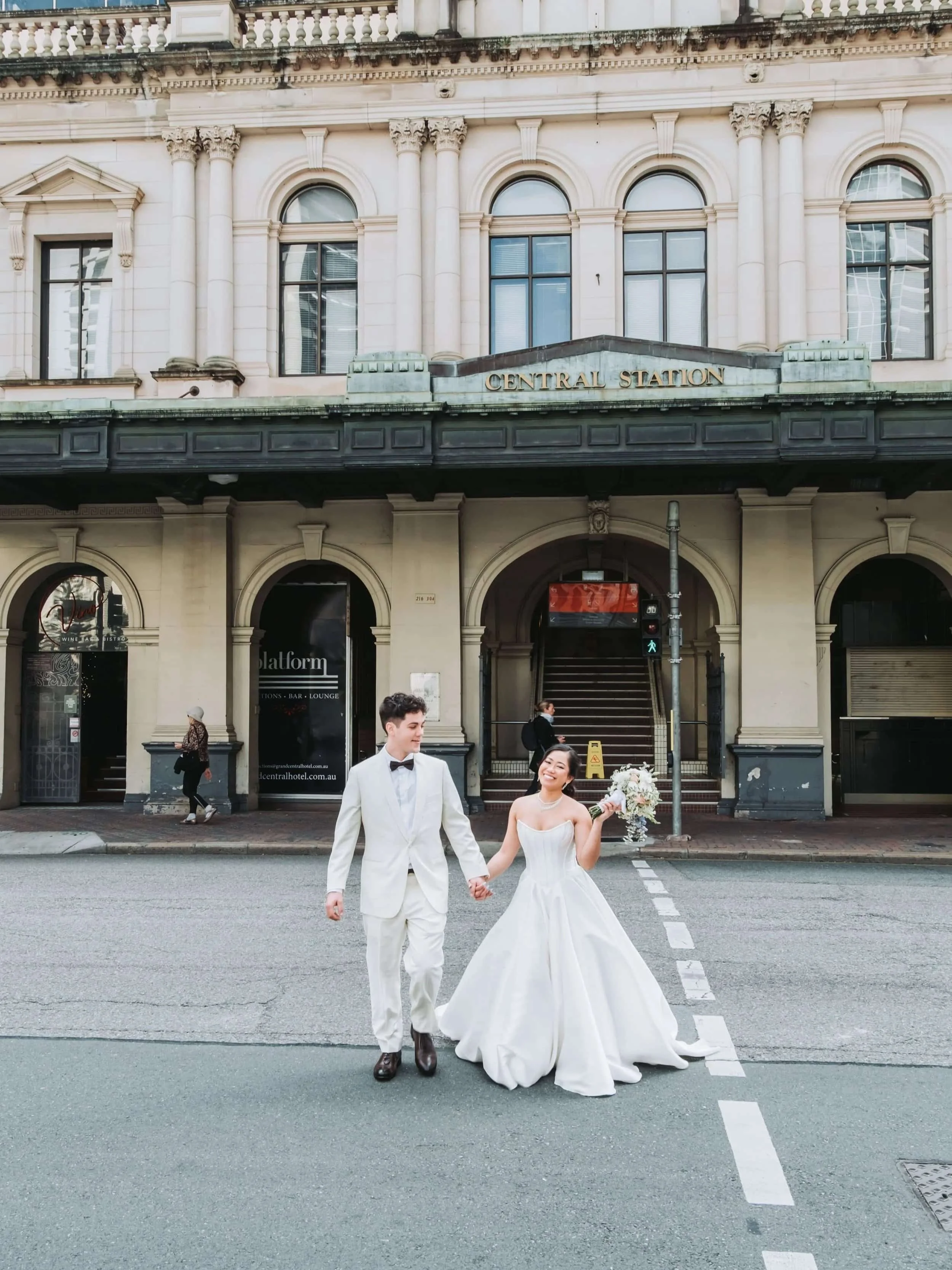 Bride and groom walk hand-in-hand across a street in front of Central Station in elegant wedding attire.