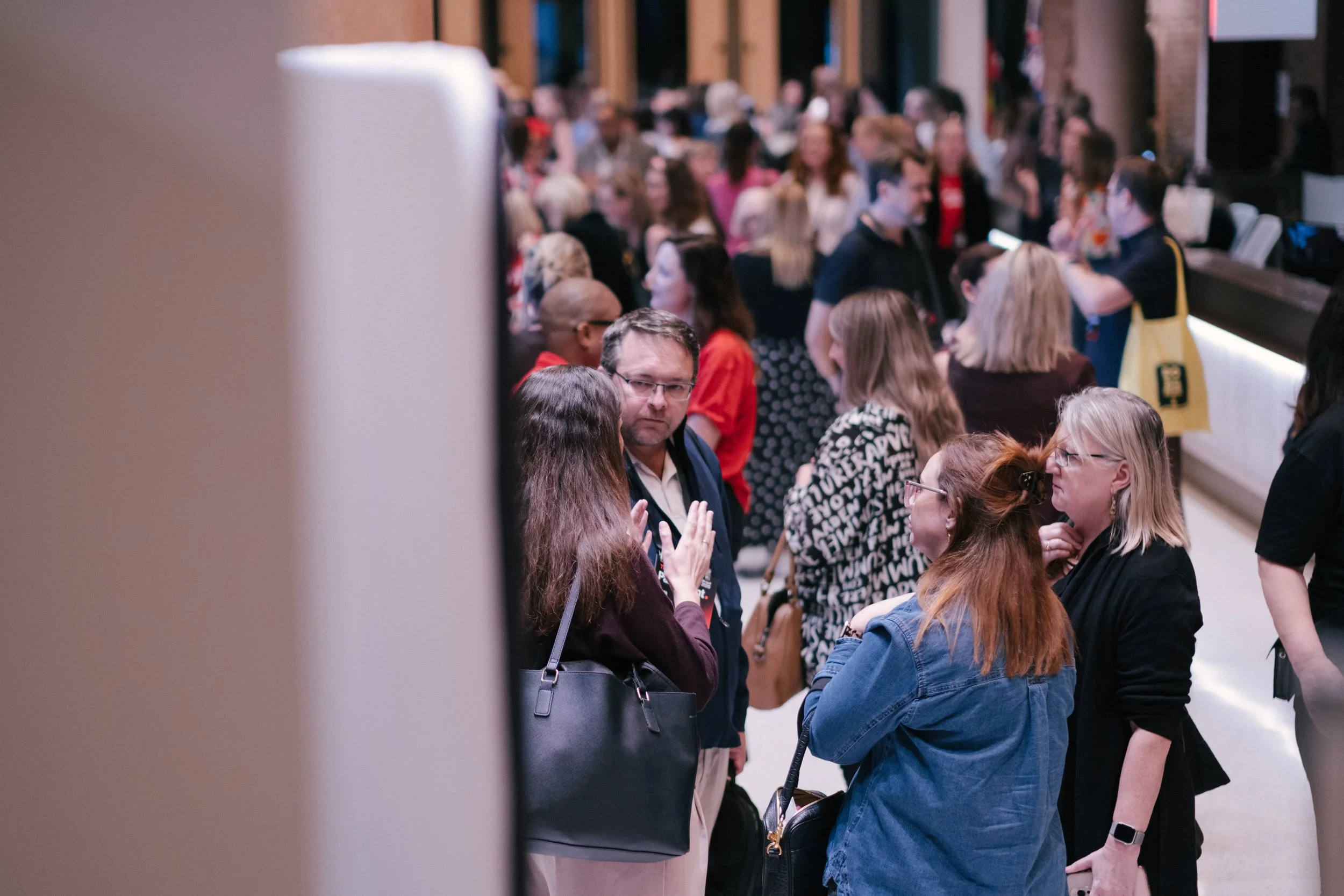 Crowd of people standing and talking in an indoor space, with some using their phones and wearing casual clothes.