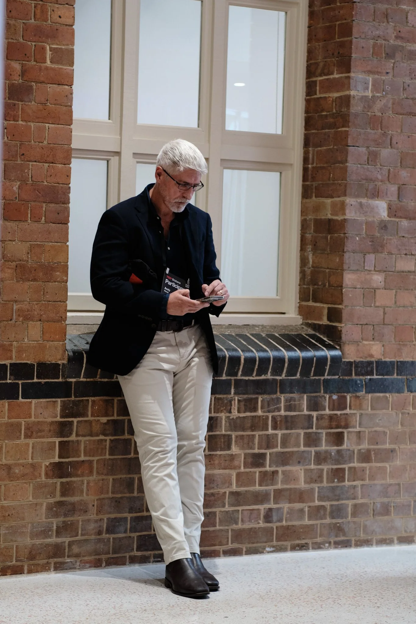 An older man with gray hair and a beard, wearing glasses, a black blazer, a name badge, light-colored pants, and black boots, is leaning against a brick wall and looking at his phone in a building with brick and glass walls.