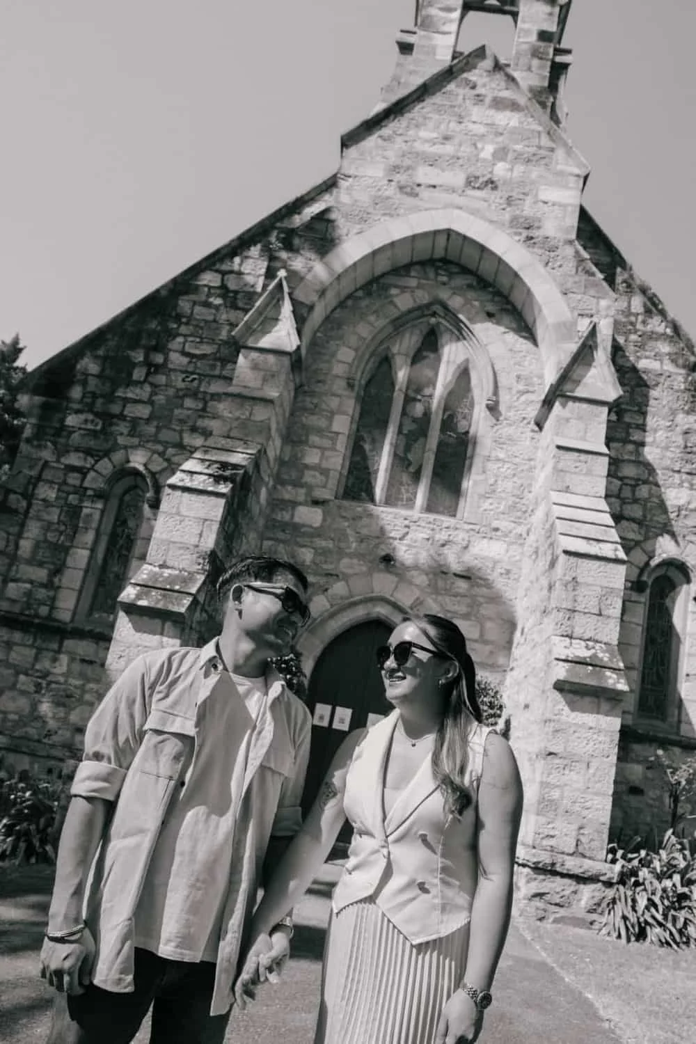 Two people, a man and a woman, wearing sunglasses and smiling, stand in front of a stone church with arched windows and ornate architecture, while holding hands.