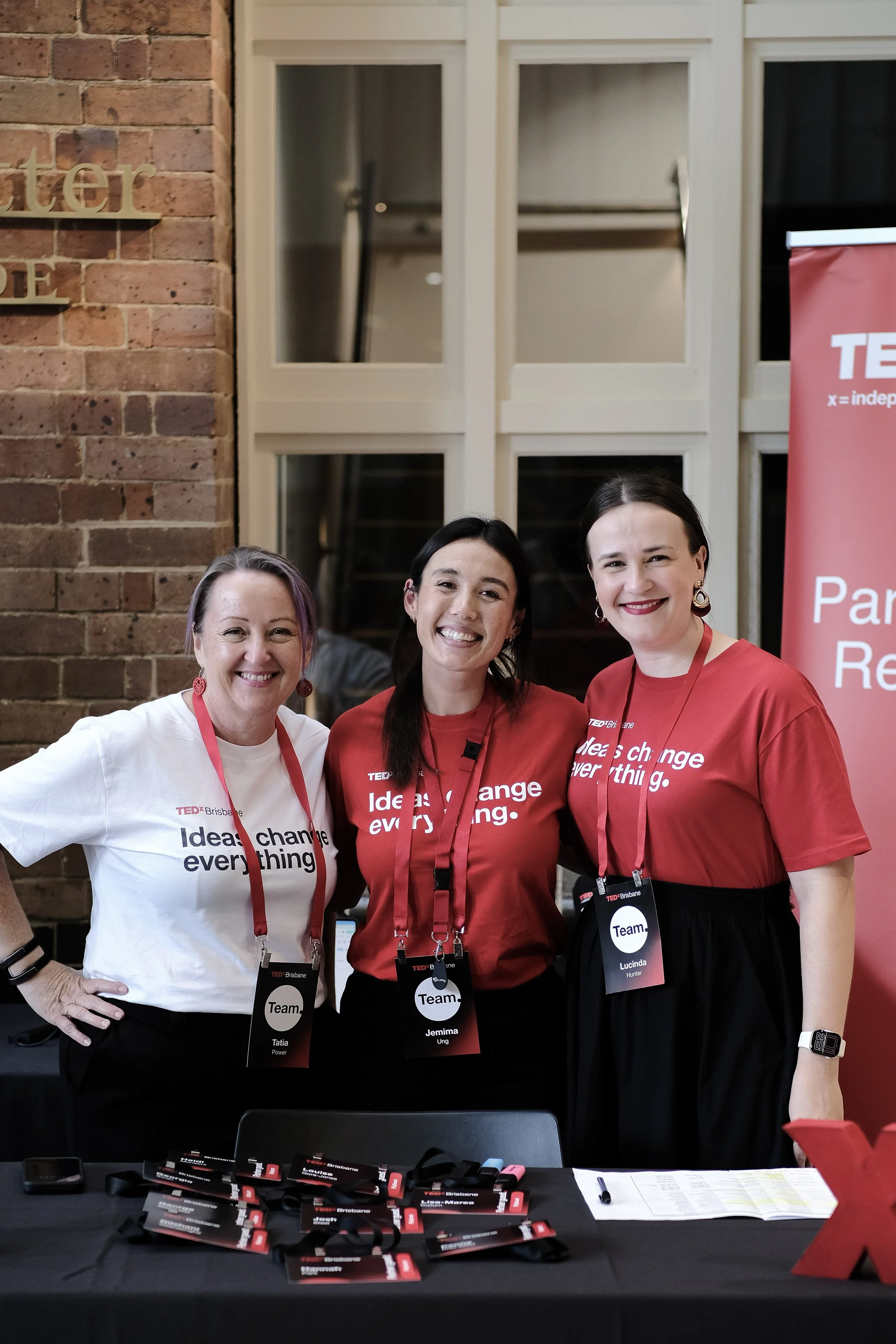 Three women in red TEDx t-shirts smile together at a TEDx event, with a TEDx banner visible behind them.