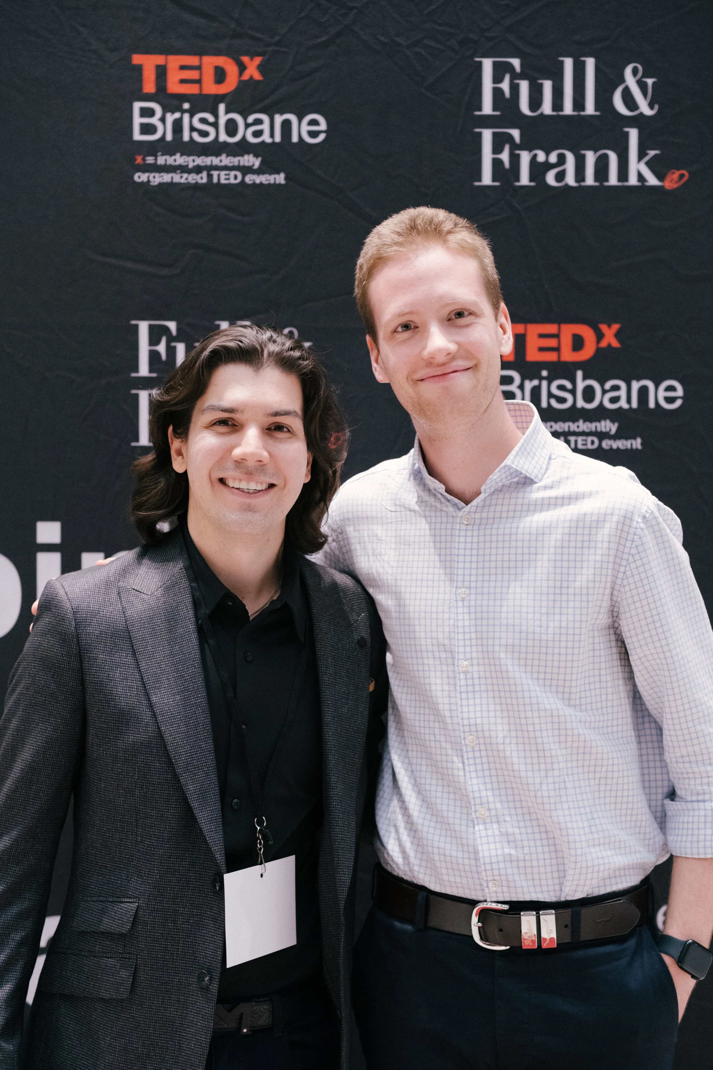 Two young men smiling at a TEDx Brisbane event with a black backdrop, one wearing a gray checkered blazer and the other in a white checkered shirt. The backdrop features the TEDx Brisbane logo and the words 'Full & Frank'.