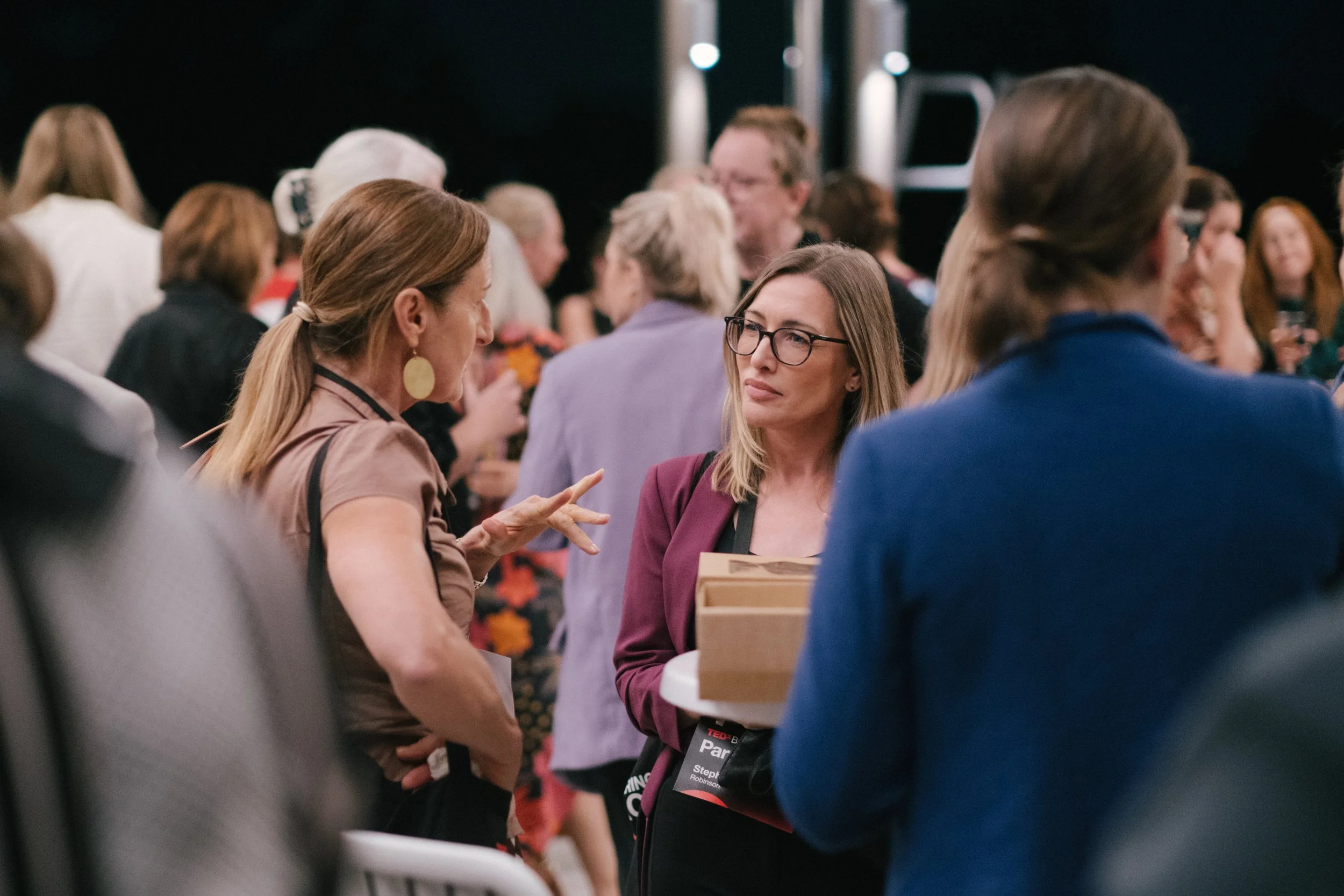 Women engaged in conversation at an indoor event, with a crowd of people in the background.