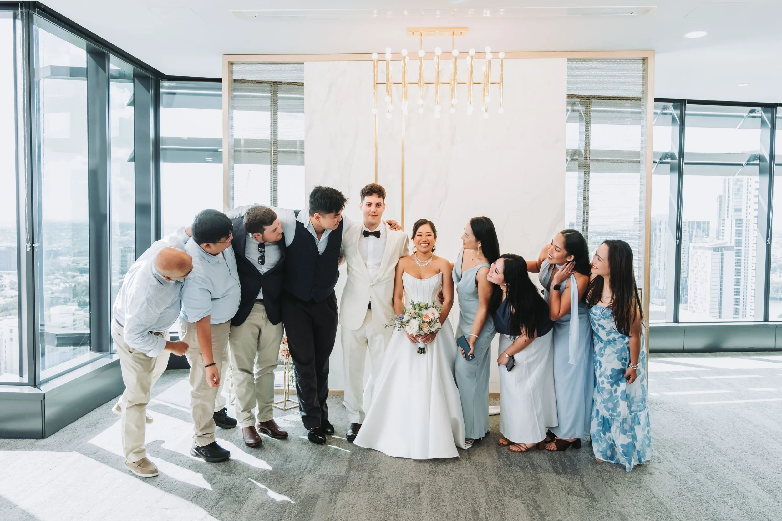 Wedding party gathers around bride and groom in elegant venue with floor-to-ceiling windows and gold chandelier backdrop.