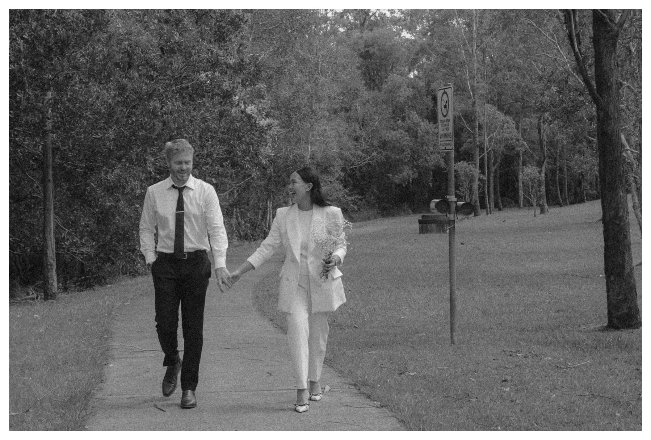 A man and woman, dressed formally, holding hands and walking outdoors on a sidewalk surrounded by trees. The woman is holding a bouquet of flowers.