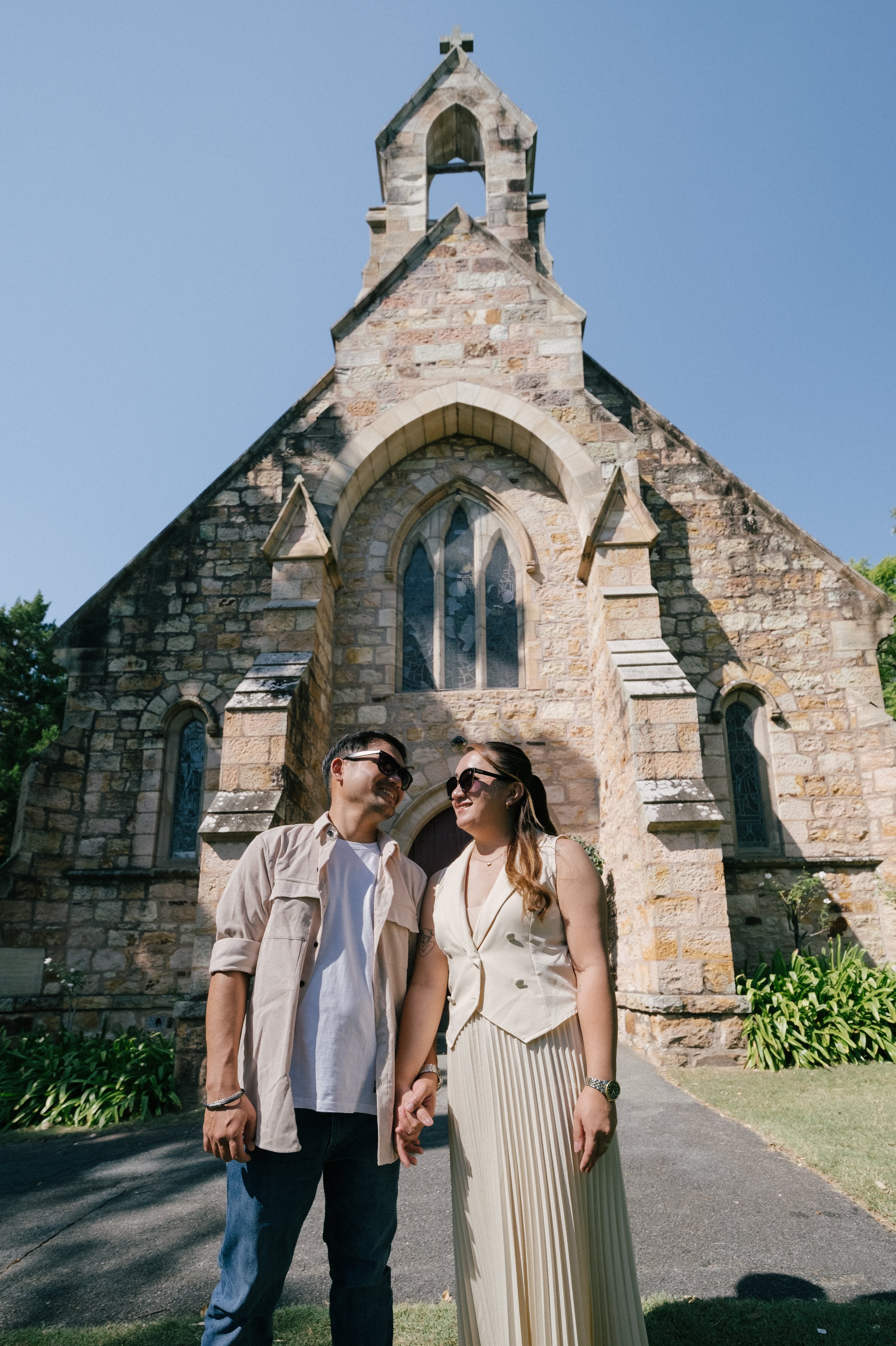 A man and a woman smiling and holding hands in front of a stone church with stained glass windows, surrounded by greenery on a sunny day.
