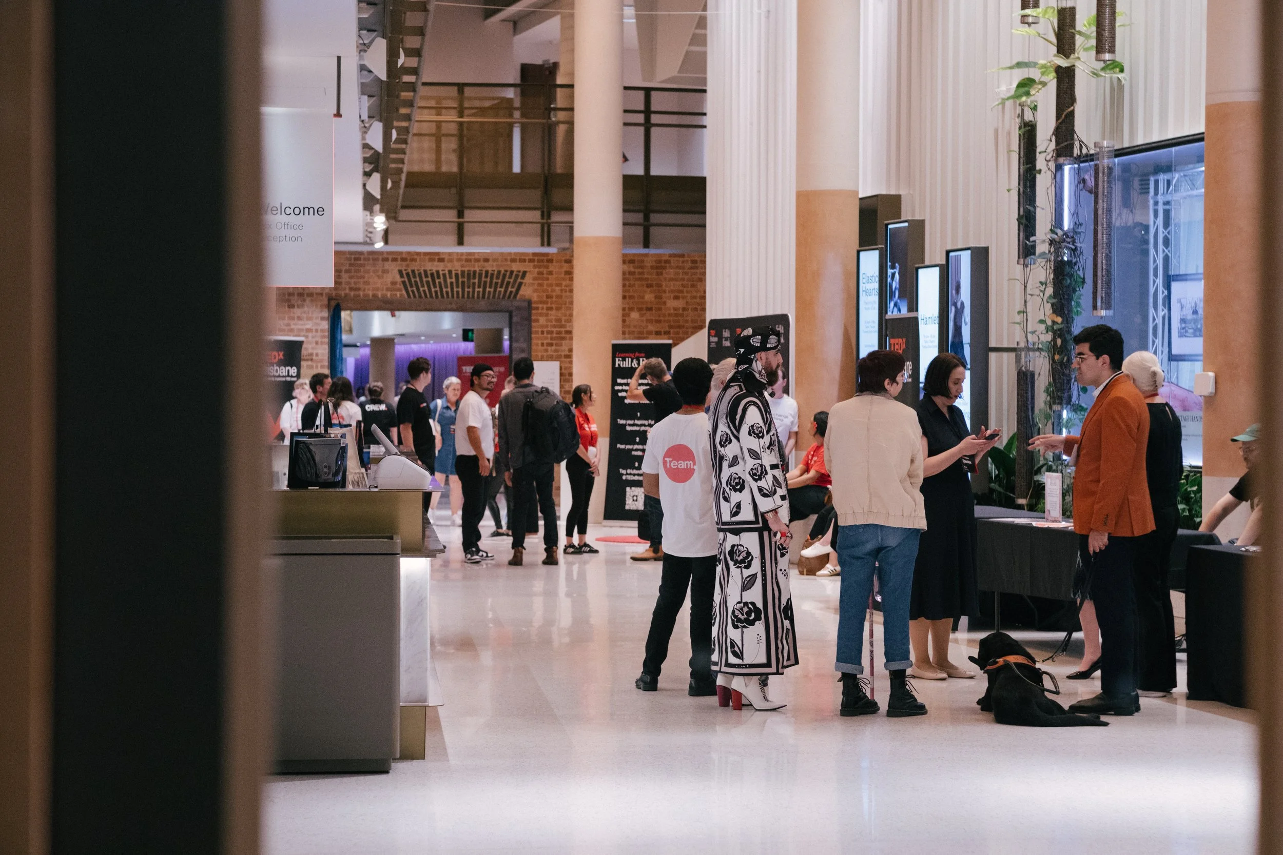 People gathering in an indoor hall for an event or conference, with large digital screens and posters visible, framed by a window or doorway.