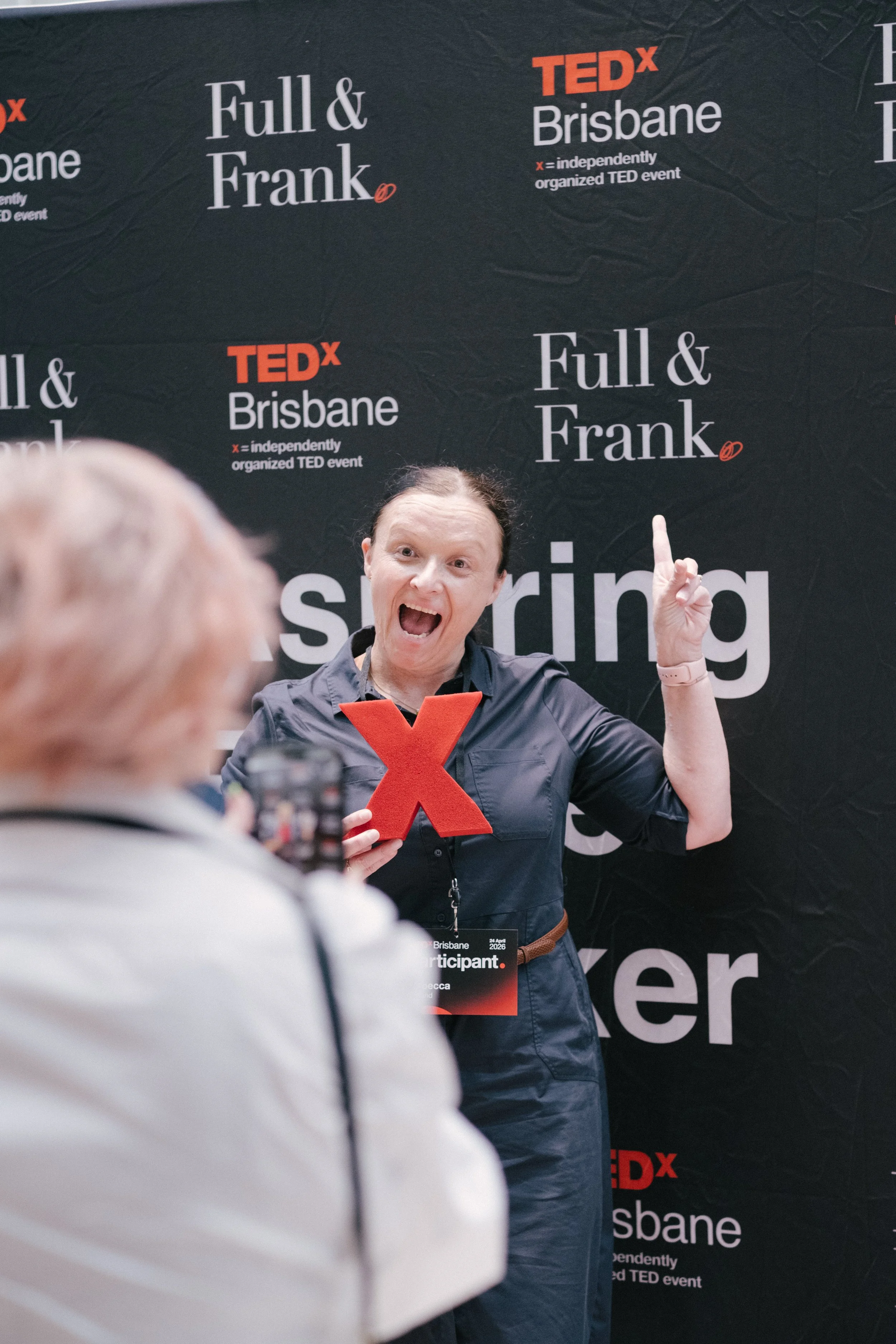 A woman at a TEDx Brisbane event holding a red X and smiling, with a large black backdrop behind her that has text including 'Full & Frank' and 'TEDx Brisbane'.
