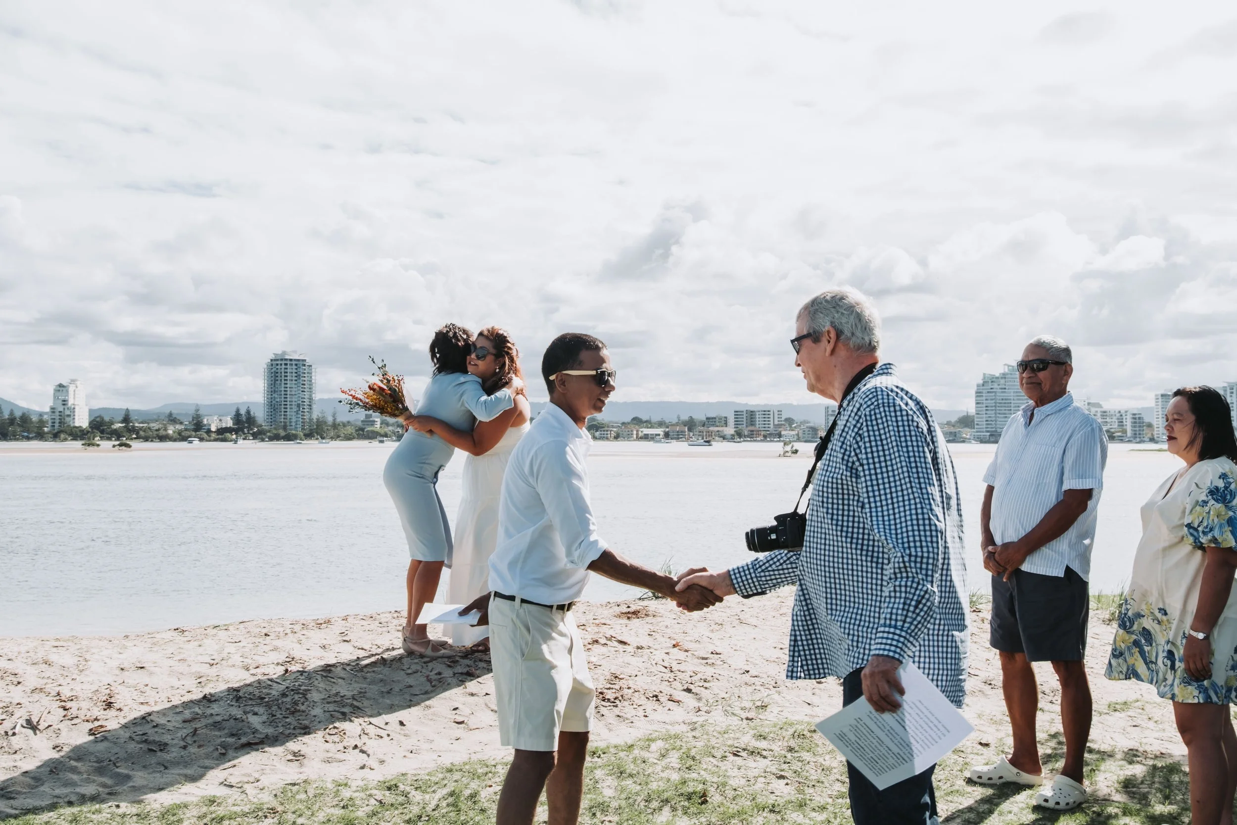 A group of people participating in a wedding or ceremony outdoors near a body of water, with buildings in the background. One woman is hugging another, and two others are engaged in conversation, all dressed in casual or semi-formal attire.